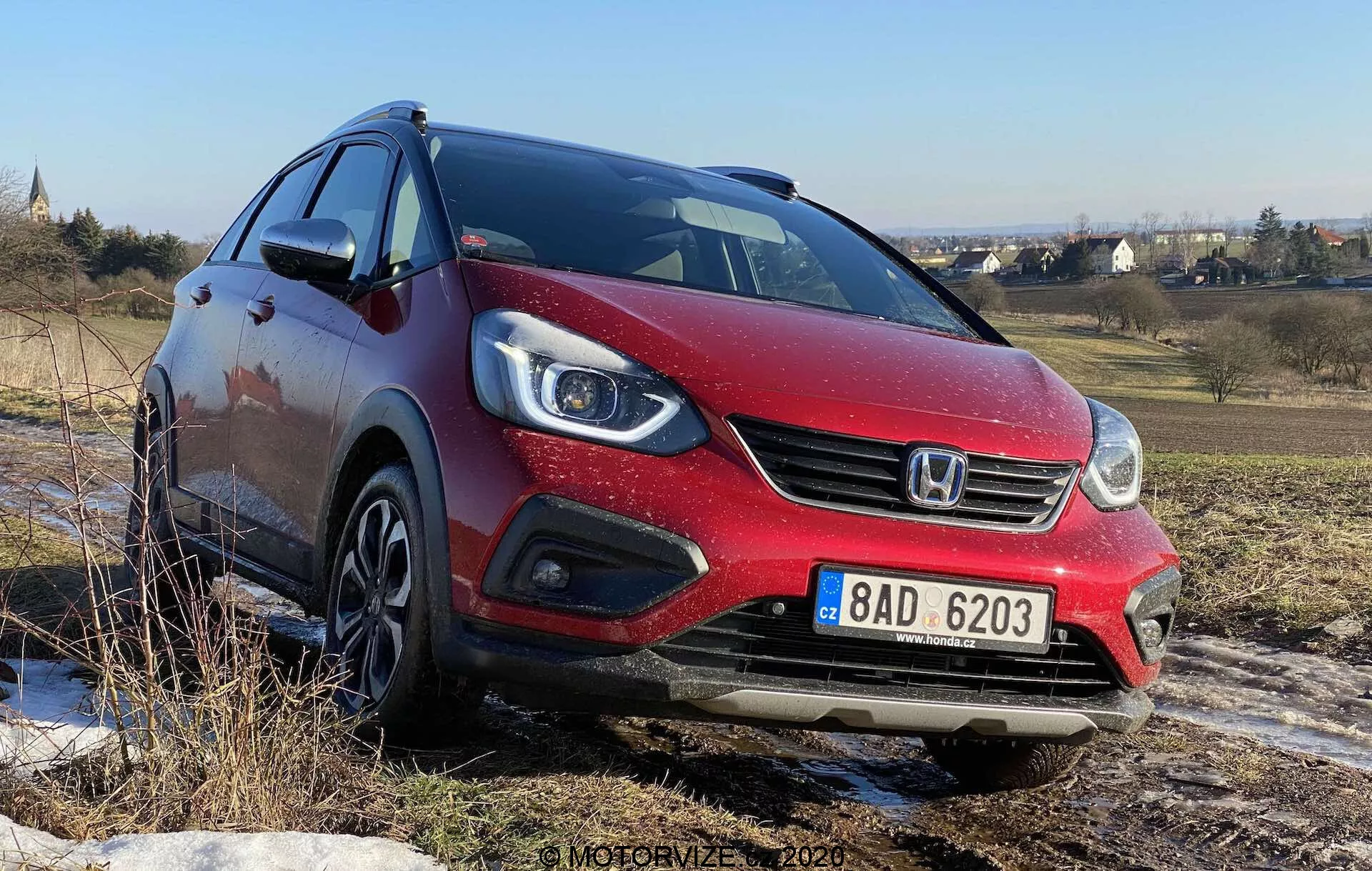 Trojpohľadový predný pohľad na červený hatchback Honda Jazz Crosstar 2020, odfotografovaný z nízkeho uhla, ktorý zobrazuje jeho prednú masku, halogénové projektorové svetlomety s integrovanými LED dennými svetlami, prednú mriežku s logom značky a zliatinové kolesá. Auto je umiestnené na nerovnom teréne pod jasnou modrou oblohou, čo naznačuje jeho schopnosť zvládať nespevnenej povrchy.