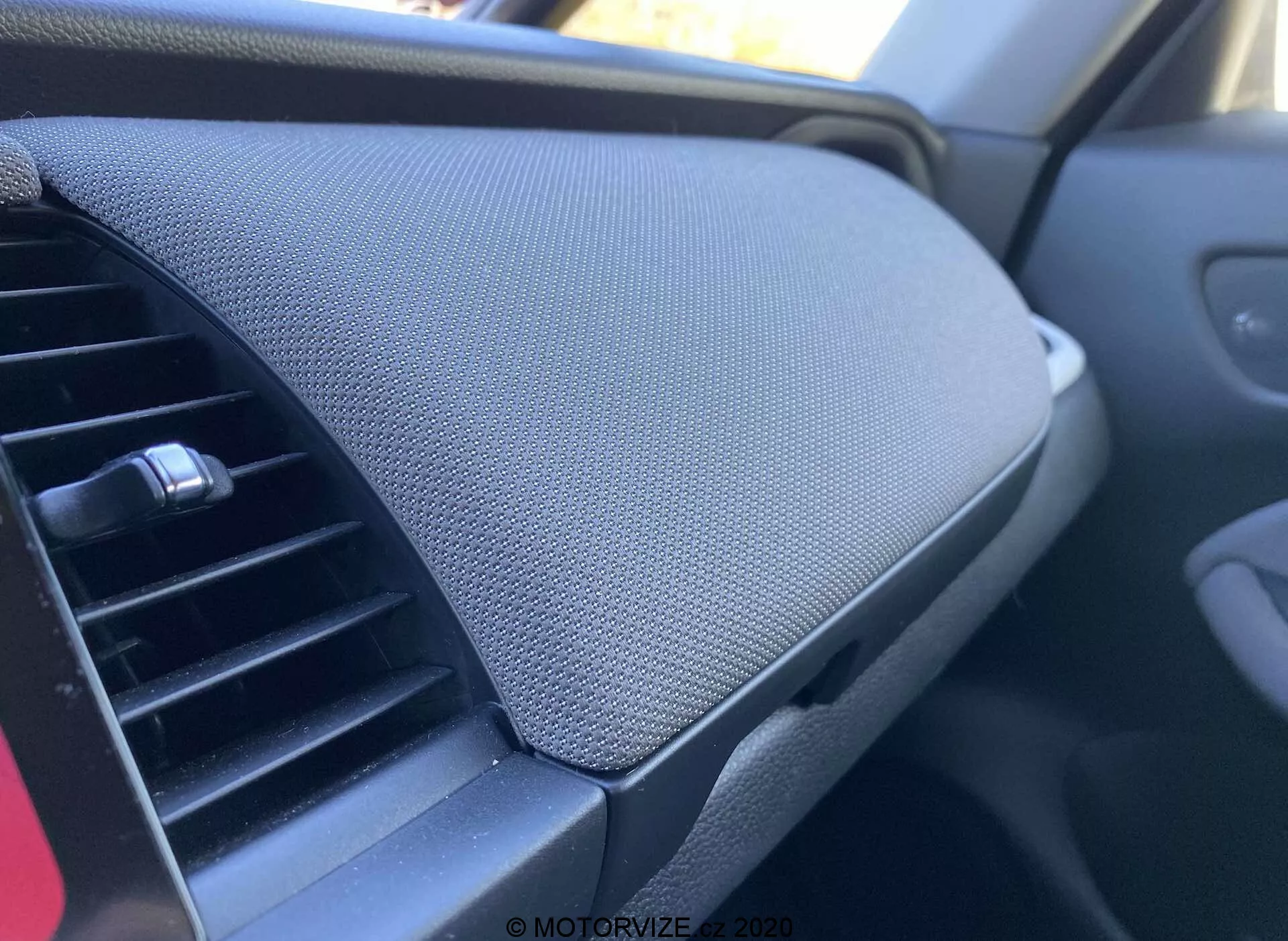 Detailný pohľad do interiéru vozidla Honda Jazz 2020 (Jazz Crosstar), zameraný na palubnú dosku. Z pohľadu spolujazdca je naľavo vidieť klimatizačný ventil s nastaviteľnou horizontálnou mriežkou. Vpravo dominuje úložný priestor palubnej dosky s uzavretým vekom charakterizovaným textúrovaným povrchom, pravdepodobne zo sieťoviny alebo látky. Priehradka je hladko integrovaná bez viditeľnej rukoväte, čo naznačuje mechanizmus otvárania na stlačenie. Celkové farebné schéma je tmavé, čo naznačuje neutrálnu estetiku zameranú na zníženie rozptýlenia.