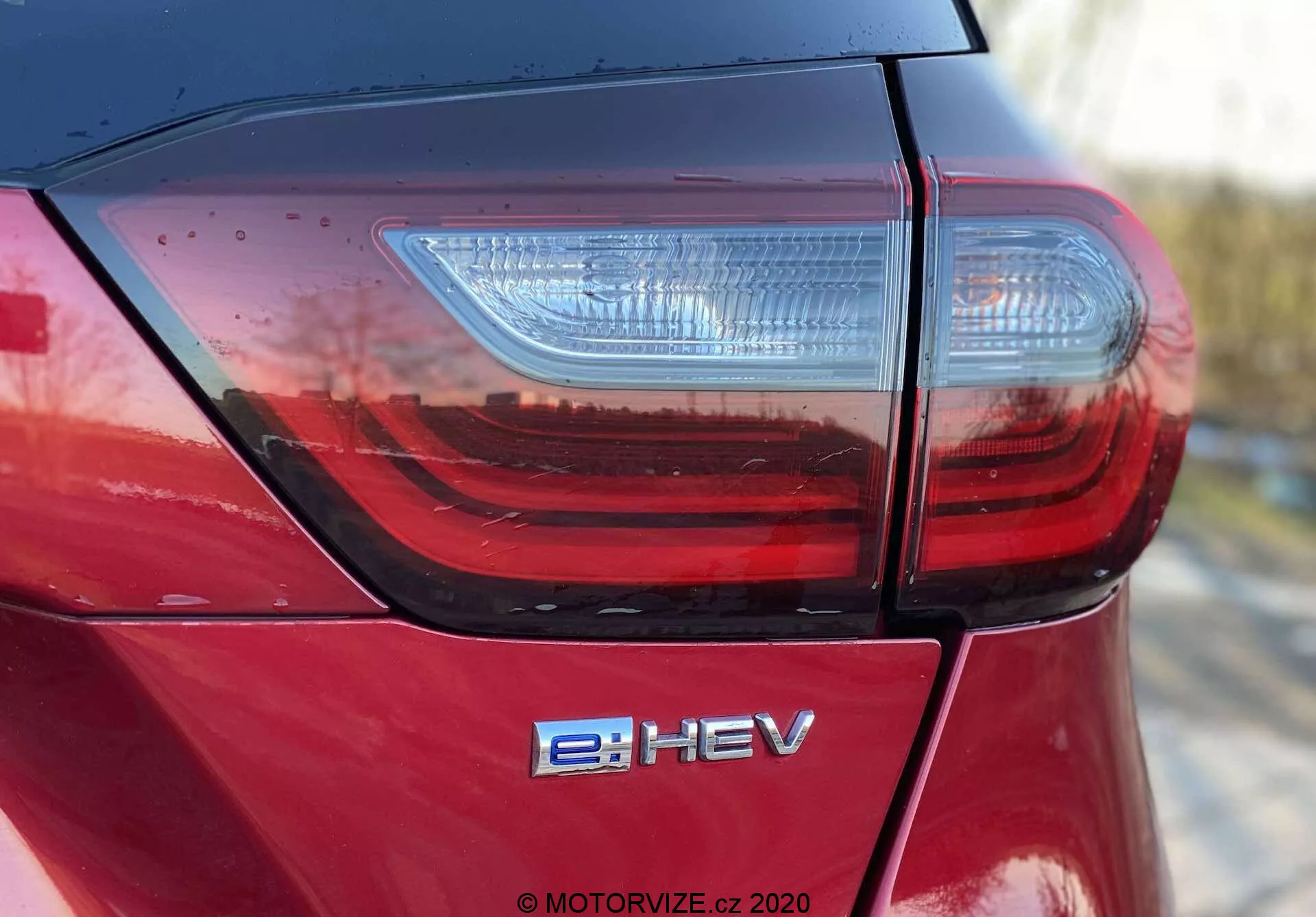 Podrobná fotografia zadnej časti Hondy Jazz Crosstar 2020 z vysokého uhla, zachytávajúca veko kufra, moderný LED zadný svetlomet a znak hybridného elektrického vozidla e:HEV. Ukazuje vysoko lesklý kovový červený lak a prémiovú kvalitu spracovania s úzkymi panelovými švami, ktoré sú zvýraznené čistými líniami a odrazmi v prirodzenom svetle.
