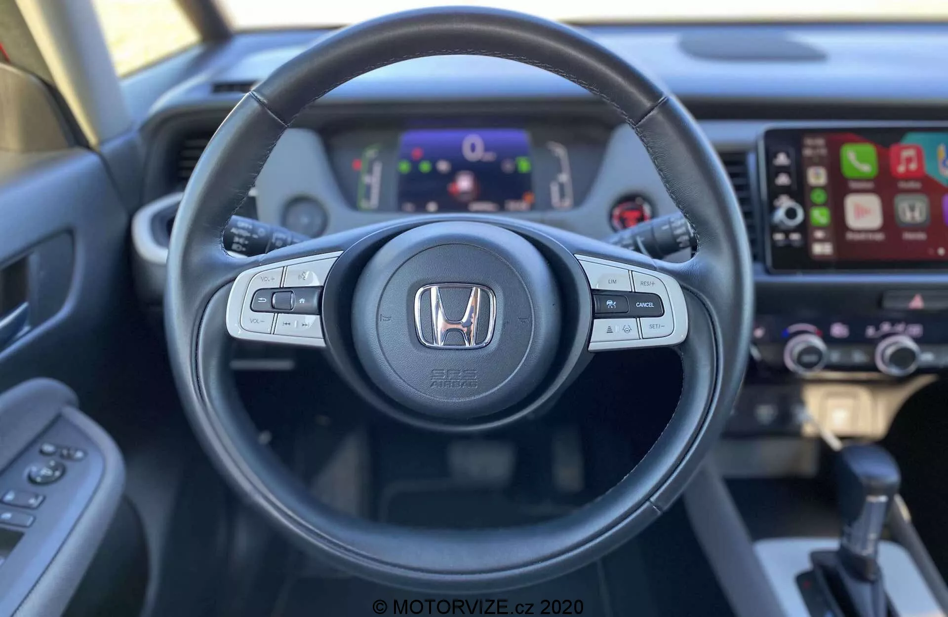 Pohľad na interiér Honda Jazz Crosstar 2020 z miesta vodiča, predstavujúci volant s ovládacími prvkami pre audio, hlasové ovládanie a tempomat. Honda znak je prominentne zobrazený uprostred. Za volantom je digitálny prístrojový panel s rôznymi ukazovateľmi a informáciami. Vpravo je viditeľný dotykový infotainment systém s kompatibilitou Apple CarPlay a mierne rozostrené ovládanie klímy. Obraz vyhotovený z pohľadu prvej osoby zdôrazňuje ergonomický dizajn a dostupnosť technológie vozidla.