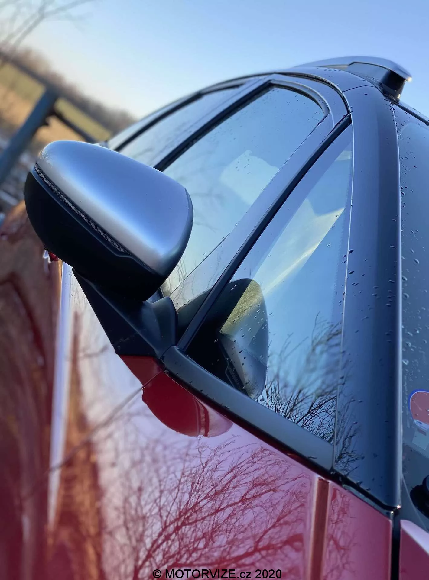 Detailný bočný pohľad na Honda Jazz 2020 (Jazz Crosstar) so zameraním na spätné zrkadlo na strane vodiča s integrovanými ukazovateľmi smeru. Obraz, zachytený z nízkeho uhla v blízkosti prednej časti vozidla, zachytáva lesklú, pravdepodobne červenú karosériu s kvapkami vody, čo naznačuje nedávny dážď alebo rannú rosu. Zrkadlo s tmavým krytom a aerodynamický dizajn vozidla sú jasne viditeľné a zdôrazňujú kvalitu vonkajšej povrchovej úpravy.