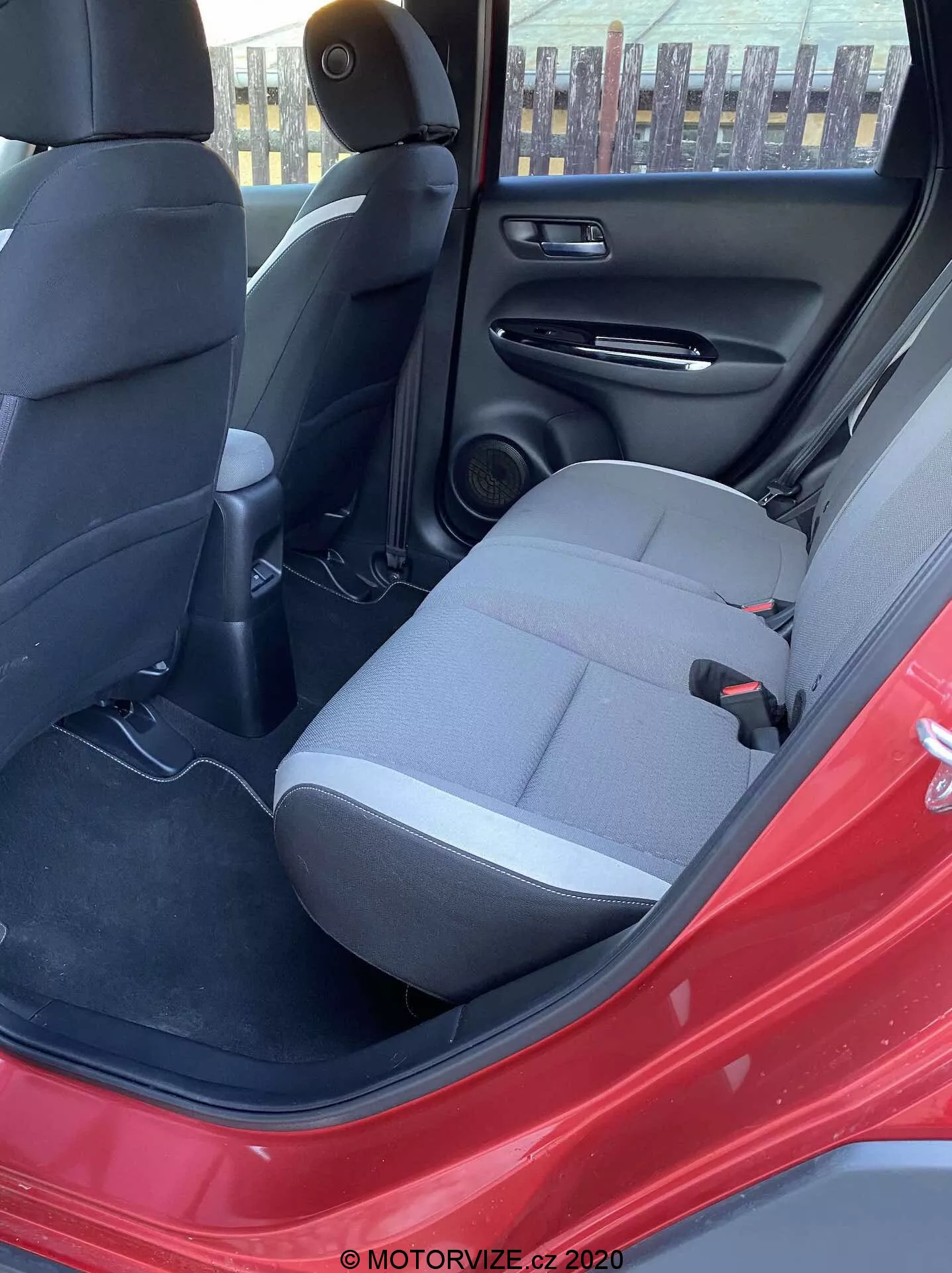 Pohľad na interiér Hondy Jazz Crosstar 2020 z otvorených zadných dverí na strane vodiča, zvýrazňujúci zadné sedadlo pre pasažiera v tmavej látke, bezpečnostný pás, zadnú stredovú konzolu s vetracími otvormi, reproduktor v zadných dverách, čierny panel dverí s odkladacím vreckom, kobercové podlahy a nápadnú červenú farbu exteriéru auta. Scénu osvetľuje prirodzené denné svetlo.