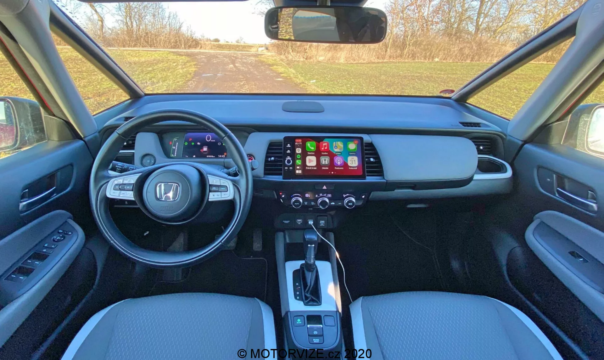 Interiér moderného auta Honda Jazz 2020 (Jazz Crosstar) pohľadom z priestoru zadných sedadiel, ukazujúci predný prístrojový panel s digitálnym prístrojovým zoskupením, veľkým infotainment dotykovým displejom, panelom ovládania klimatizácie, automatickou radiacou pákou, koženými sedadlami a ovládacími prvkami na dverách.