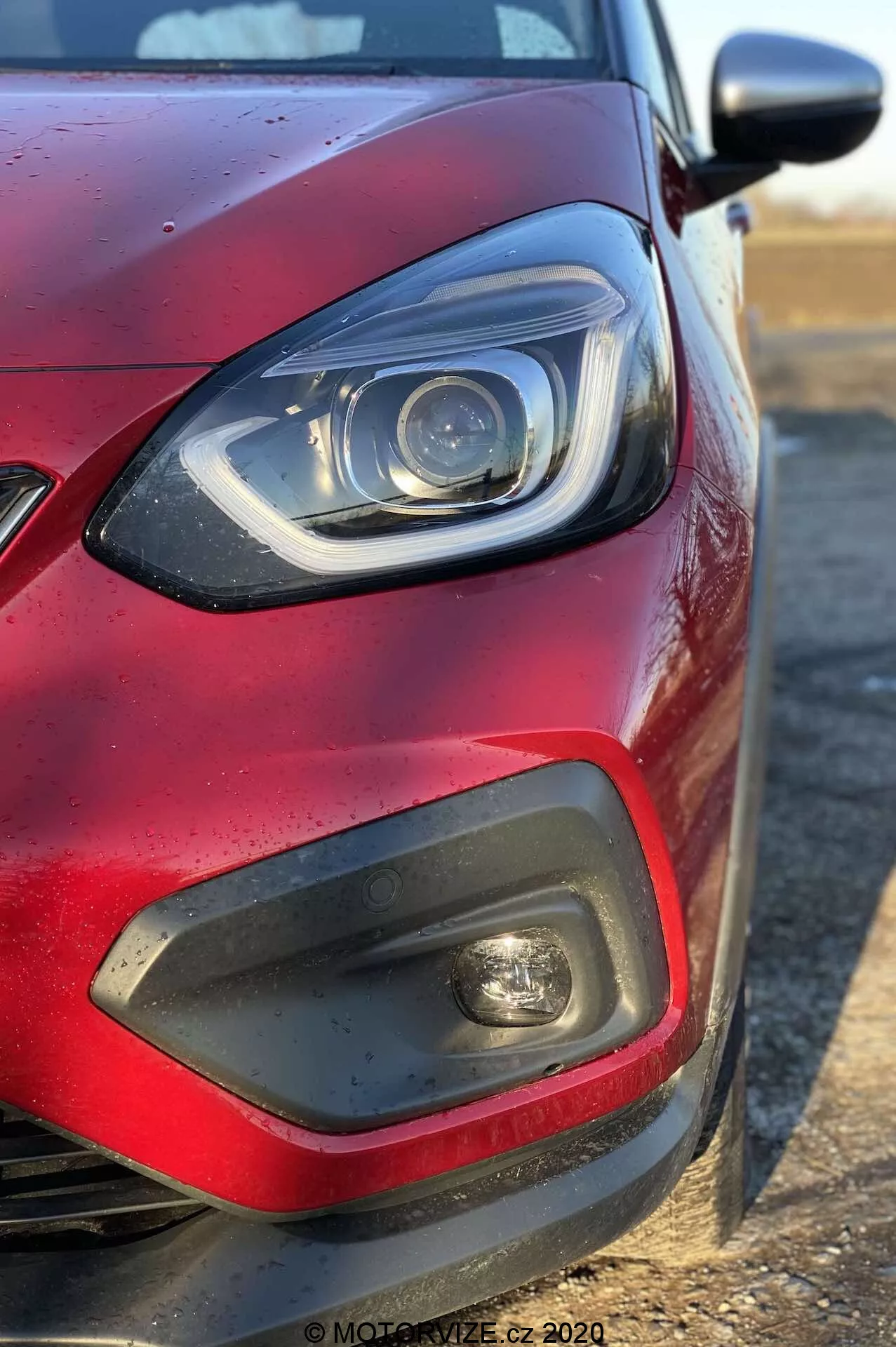 Detailný záber na predný ľavý kvadrant červeného Honda Jazz 2020 (Jazz-Crosstar) z bočného uhla, sústrediaci sa na svetlomet s LED alebo xenónovými prvkami a zakrivenou DRL lištou, čierny plastový inzert s hmlovým svetlom, časť mriežky, metalickú červenú farbu s kvapkami vody a telo farebné bočné zrkadlo vyčnievajúce z pravej strany.