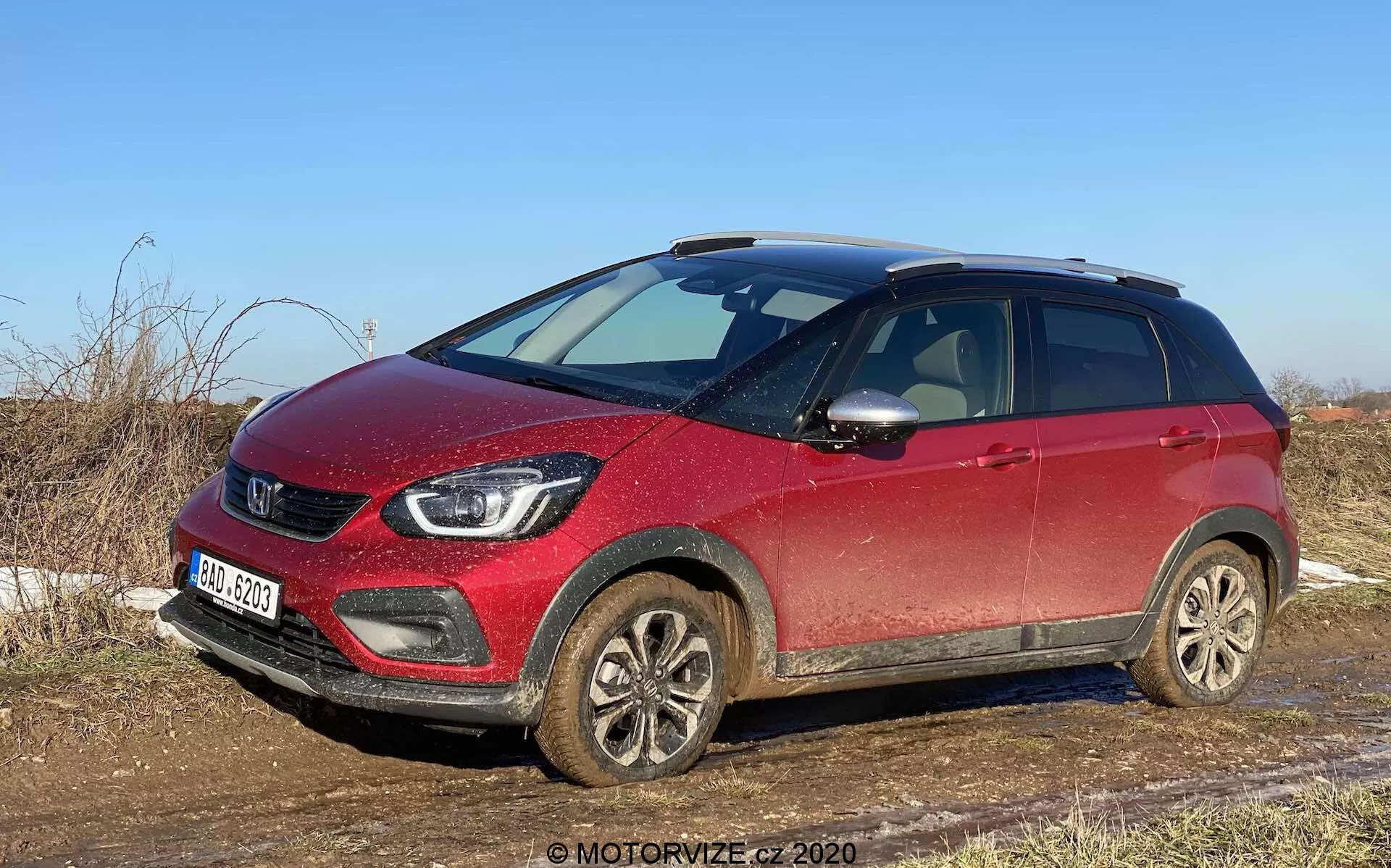 Trošku zošikmený predný pohľad na červené kompaktné crossover SUV Honda Jazz Crosstar 2020, zachytené za denného svetla z pohľadu spolujazdca, ukazujúci prednú mriežku, svetlomety, zablatené zliatinové kolesá a bočný profil až po zadné dvere pre pasažierov. Auto vyzerá mierne špinavé, čo naznačuje nedávne použitie na nespevnených alebo mokrých cestách. Viditeľné vlastnosti zahŕňajú výraznú mriežku so značkovým logom, moderné LED svetlomety, hmlové svetlá, zliatinové kolesá, zrkadlá s integrovanými smerovkami, strešné nosiče a zvýšenú svetlú výšku, čo naznačuje všestrannosť a schopnosť vozidla na ľahký terén. SUV je zaparkované na vidieku so suchou trávou a čistým nebom, zdôrazňujúc jeho vhodnosť na vidiecke cesty a ľahký terén.