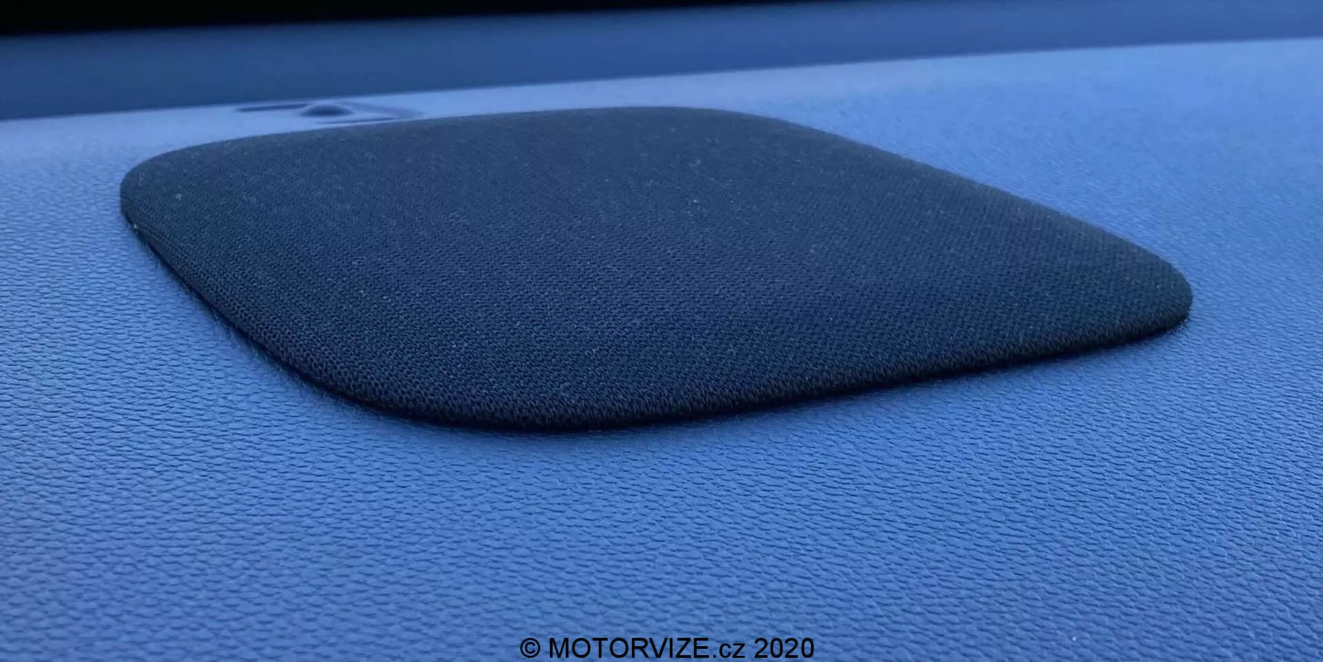 Detailná fotografia mriežky reproduktora v aute, zabudovanej do tmavomodrého alebo čierneho palubného panela alebo dverového panelu, pravdepodobne Honda Jazz 2020. Mriežka reproduktora je vyrobená z textúrovaného materiálu podobného látke a má tvar skráteného lichobežníka alebo nepravidelného štvoruholníka s mierne konvexnou stranou. Komponent je mierne vyvýšený a vrhá tieň, čo zdôrazňuje jeho profil a textúru. Fotografia je urobená bočným uhlom za použitia rozptýleného prirodzeného svetla, zvýrazňuje zrnitú textúru okolitého povrchu a obsahuje vodoznak s nápisom '© MOTORVIZE.cz 2020.'