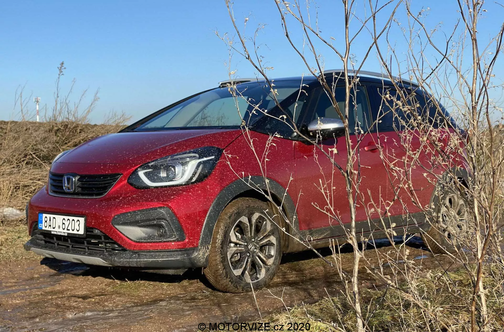 Denná vonkajšia fotografia Honda Jazz 2020 (Jazz Crosstar) zaparkovaného na trávnatom teréne. Obrázok zachytáva subkompaktné SUV s výraznou červenou farbou, výraznou mriežkou chladiča a modernými svetlometmi s LED osvetlením. Auto je zobrazené z trojštvrťového predného uhla ukazujúceho zvýšenú svetlú výšku, hliníkové kolesá, čierne plastové obloženie okolo oblúkov kolies a spodnej časti karosérie. Jasná modrá obloha a prirodzené osvetlenie a okolité konáre čiastočne zakrývajú spodnú prednú časť. Viditeľné sú bočné zrkadlá s integrovanými smerovkami a pohľad dovnútra vozidla cez čisté okno vodiča.
