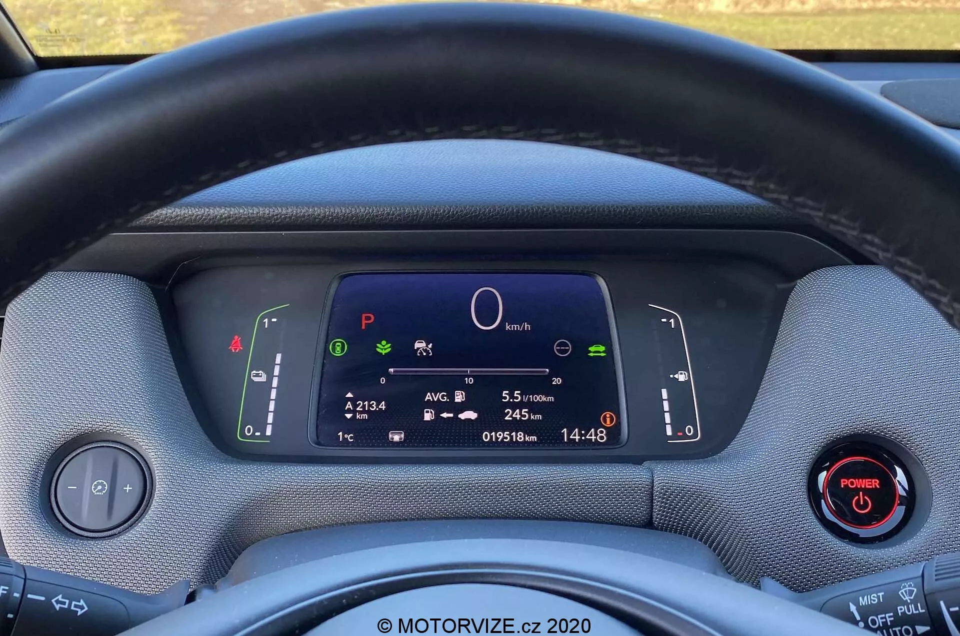 Detailný záber interiéru Honda Jazz Crosstar 2020 z pohľadu vodiča, ukazujúci volant a plne digitálny prístrojový panel. Textilný poťah volantu čiastočne zakrýva pohľad na digitálny displej, ktorý zobrazuje symboly systémov vozidla, centrálny kruhový graf, informácie o ceste, odometer a hodiny. Za volantom sú viditeľné dve páčky ovládajúce smerovky, svetlá a stierače. Na ľavej strane je ovládací panel pre svetlomety a na pravej strane tlačidlo s červeným krúžkom. Materiály palubnej dosky majú textúrovaný vzhľad, čo zvýrazňuje ekologický dizajn. Obrázok zdôrazňuje moderné technológie, digitálny prístrojový panel a potenciálne funkcie hybridného alebo elektrického vozidla.