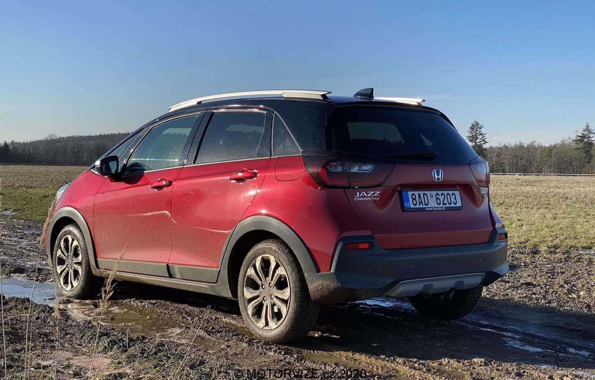 Pohľad zo zadnej tretiny na červený Honda Jazz Crosstar 2020, ktorý ukazuje jeho vlastnosti subkompaktného crossover SUV. Obrázok nasnímaný z nízkeho uhla zvýrazňuje výšku vozidla a svetlú výšku, s viditeľnými zadnými svetelnými zhlukmi, označením 'JAZZ', liatinovými kolesami, systémom strešných líšt, tónovanými zadnými oknami, blatom a viditeľnou poznávacou značkou, zaparkovaným na blatistom poli.