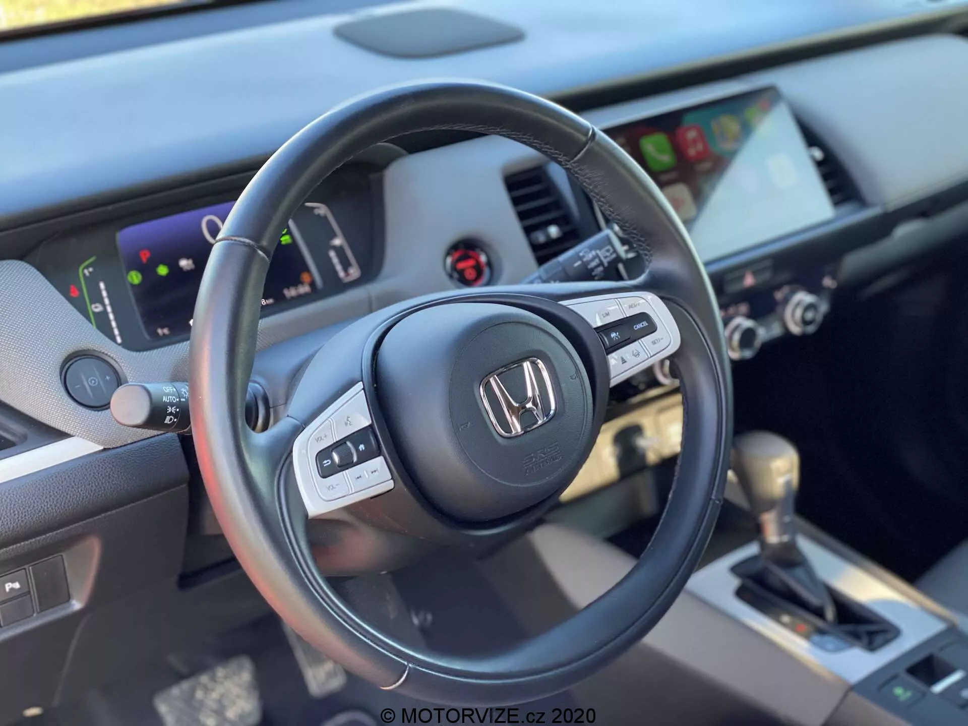 Detailný pohľad na interiér Honda Jazz Crosstar 2020 z pohľadu sedadla vodiča, ktorý zvýrazňuje ľavú stranu koženého trojramenného volantu s multifunkčnými ovládacími prvkami a prístrojovú dosku s digitálnym prístrojovým panelom a dotykovým infotainment systémom. Obrázok zahŕňa ovládače klimatizácie a automatickú radiacu páku s koženým poťahom na stredovej konzole, zdôrazňujúc moderný a dobre vybavený dizajn.