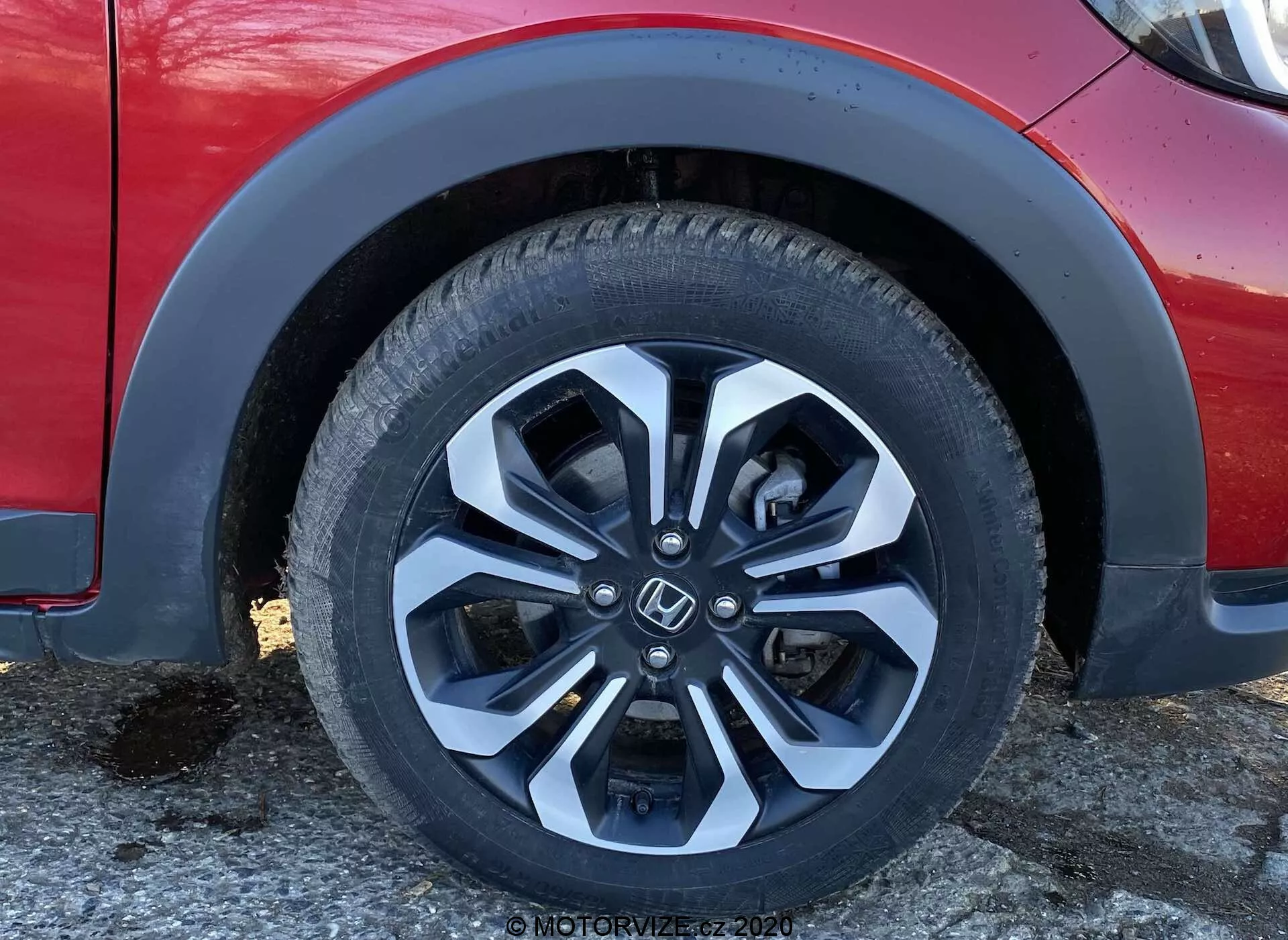 Detailný záber na prednú ľavú koleso a spodnú časť karosérie Honda Jazz Crosstar 2020, zdôrazňujúci moderný, viacpaprskový hliníkový disk s dvojtónovou povrchovou úpravou (strieborno-čiernou), detaily dezénu pneumatík a informácie o bočnici. Obrázok tiež ukazuje oblúk blatníka, časť predného nárazníka a sekciu bočných dverí z mierne nízkeho uhla, zdôrazňujúc dizajnové a technologické aspekty, ako je technológia pneumatík, dizajn diskov, zloženie materiálov, aerodynamika a náznaky odpruženia vozidla.