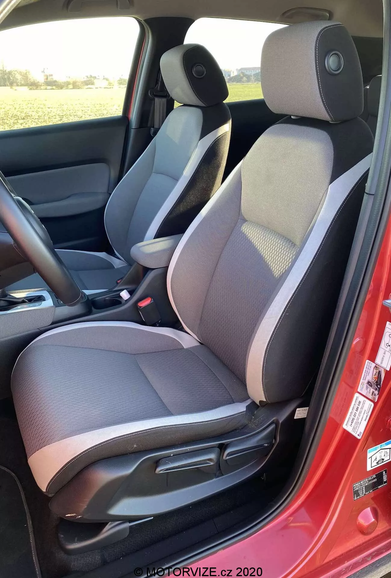 Interiér vozidla Honda Jazz Crosstar 2020 z ľavej strany, zvýrazňujúci dvojfarebné čalúnené sedadlo vodiča so vzorovanou strednou časťou, nastaviteľnú opierku hlavy, ručné nastavenie sedadla, červený bezpečnostný pás, reproduktor na paneli dverí, tmavú podlahovú rohož, stredovú konzolu s lakťovou opierkou, ovládanie bočného zrkadla a tmavo-svetlú farebnú schému.