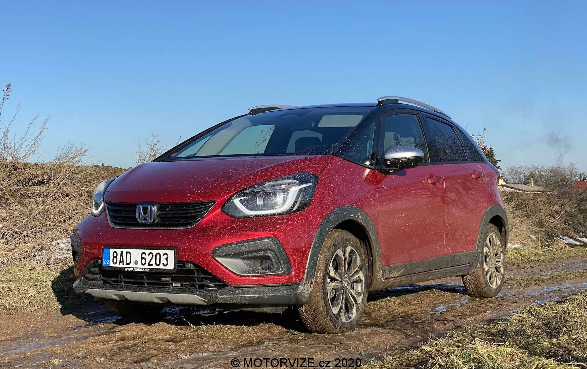 Pohľad z predného uhla na červený Honda Jazz Crosstar 2020, kompaktné crossover SUV, zachytené na terénnom povrchu s viditeľným blatom a špinou. Obrázok zdôrazňuje prednú mriežku, elegantné svetlomety, ktoré môžu obsahovať LED alebo projektorové lampy, hmlové svetlá integrované do nárazníka a jemne sklonenú kapotu. Vozidlo, vybavené strešnými nosičmi, kolesami pokrytými blatom a bočnými zrkadlami s integrovanými smerovkami, naznačuje moderné bezpečnostné funkcie a pokročilé technologické vybavenie. Ideálny pre kategórie zamerané na vozidlá, recenzie áut, terénne schopnosti, automobilovú techniku a dizajn.