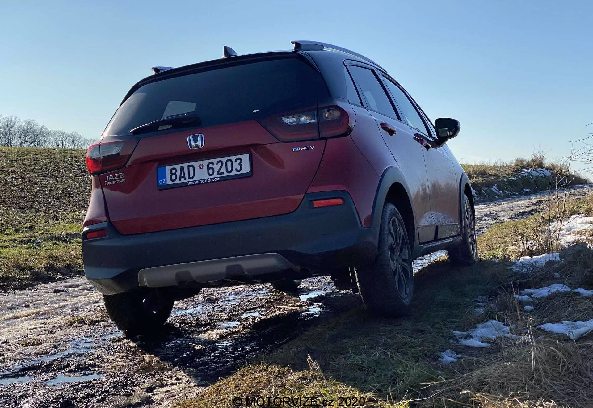 Zadný pohľad z troch štvrtín na červené SUV Honda Jazz Crosstar, ktorý ukazuje dizajn zadných svetiel, zadného nárazníka, výfukového systému a zliatinových kolies. Uhol zdôrazňuje svetlú výšku vozidla a zdá sa, že nedávno prechádzalo zasneženými a blatistými cestami, čo naznačuje jeho všestrannosť v rôznych podmienkach.