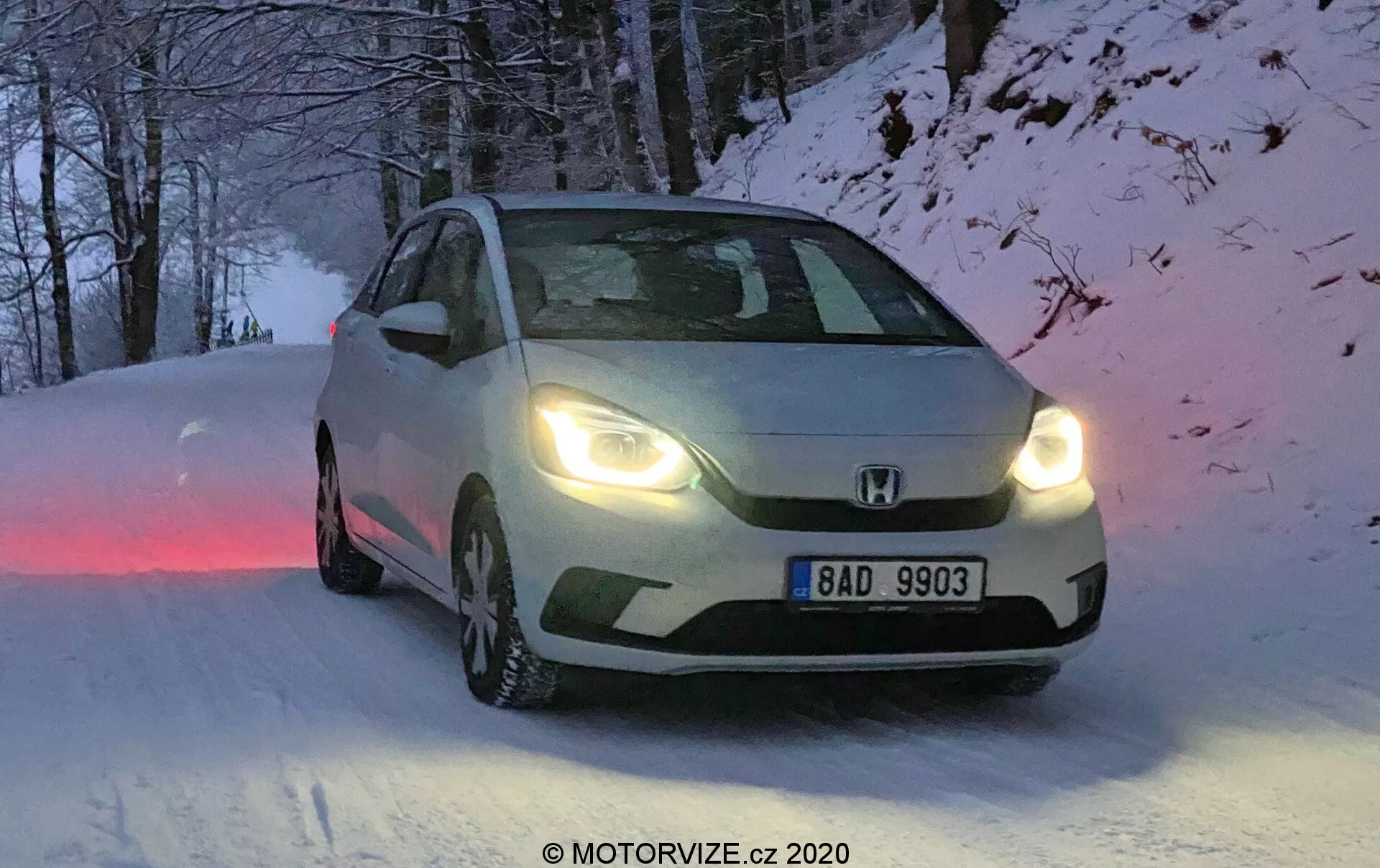 Tento obrázok zachytáva Honda Jazz 2020 z trojštvrťového predného uhla, zobrazujúc dizajnové prvky prednej mriežky, svetlometov a kontúr kapoty. Vozidlo, ktoré je umiestnené na zasneženej ceste, má zimné pneumatiky s viditeľným vzorom dezénu a zapnuté svetlomety, čo naznačuje svitanie, súmrak alebo zníženú viditeľnosť. Európska registračná značka má nápis „BAD 9903“, a na obrázku je mierne rozmazanie pohybu, ktoré pridáva dynamiku. Prirodzené, chladné osvetlenie a zimné prostredie dopĺňajú schopnosti vozidla do všetkých podmienok.