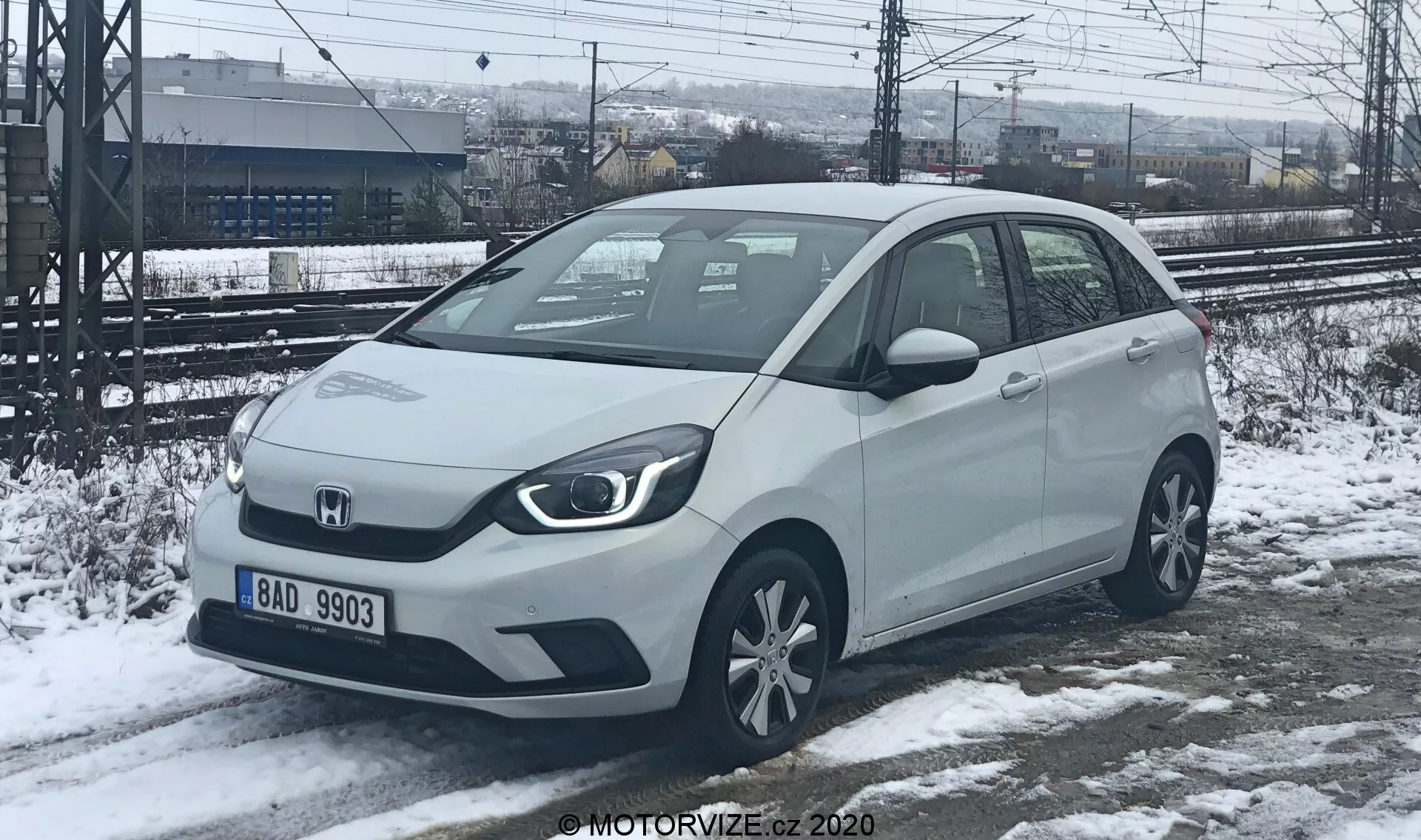 Pohľad spredu z troch štvrtín na kompaktné auto Honda Jazz 2020 vo svetlej farbe, pravdepodobne bielej alebo striebornej, zachytené v zimnom prostredí so snehom na zemi. Ostré, uhlové svetlá a pevná mriežka naznačujú možnosti elektrifikovaného pohonu. Auto má viditeľné parkovacie senzory na prednom nárazníku, zrkadlá namontované na dverách s integrovanými smerovkami a štandardné zliatinové kolesá. Zaparkované na čiastočne roztopenom snehu, s koľajnicami a plotom v pozadí, naznačujúce priemyselnú alebo železničnú stanicu.