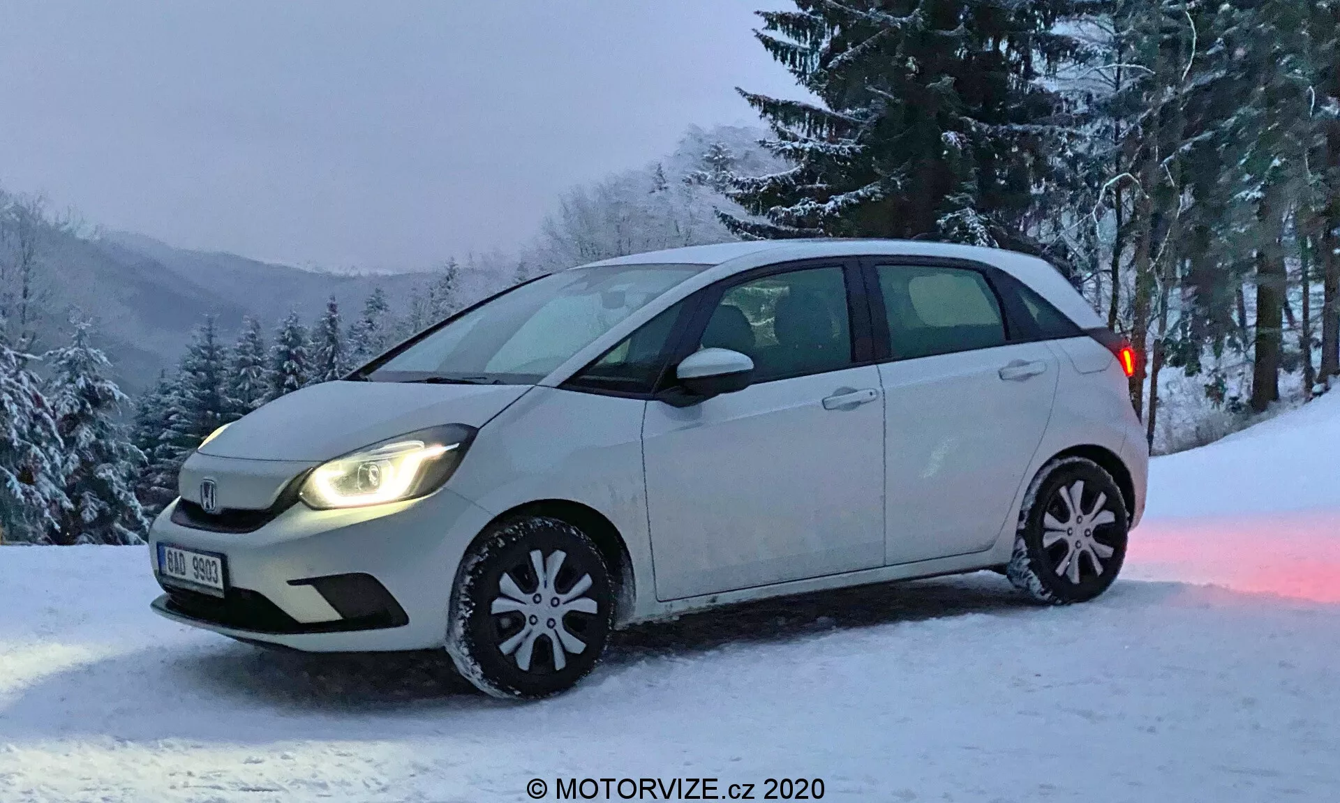 Hatchback Honda Jazz z roku 2020 fotografovaný zo šikmého predného uhla, ktorý zachytáva celý pohľad na stranu vodiča a čiastočne ukazuje predný nárazník a mriežku chladiča. Vozidlo má dvojfarebný lak, väčšina auta je v svetlej farbe, spodná časť dverí je tmavšia. Svetlomety sú zapnuté so štandardnými halogénovými lampami, bez hmlových svetiel. Dekoratívne zimné kryty kolies so snehovými vločkami naznačujú zimnú sezónu. Auto je zaparkované na zasneženom mieste s pozadím stromov a zasneženého kopca, čo naznačuje vidiecku alebo horskú oblasť. Obrázok s láskavým dovolením MOTORVIZE.cz.