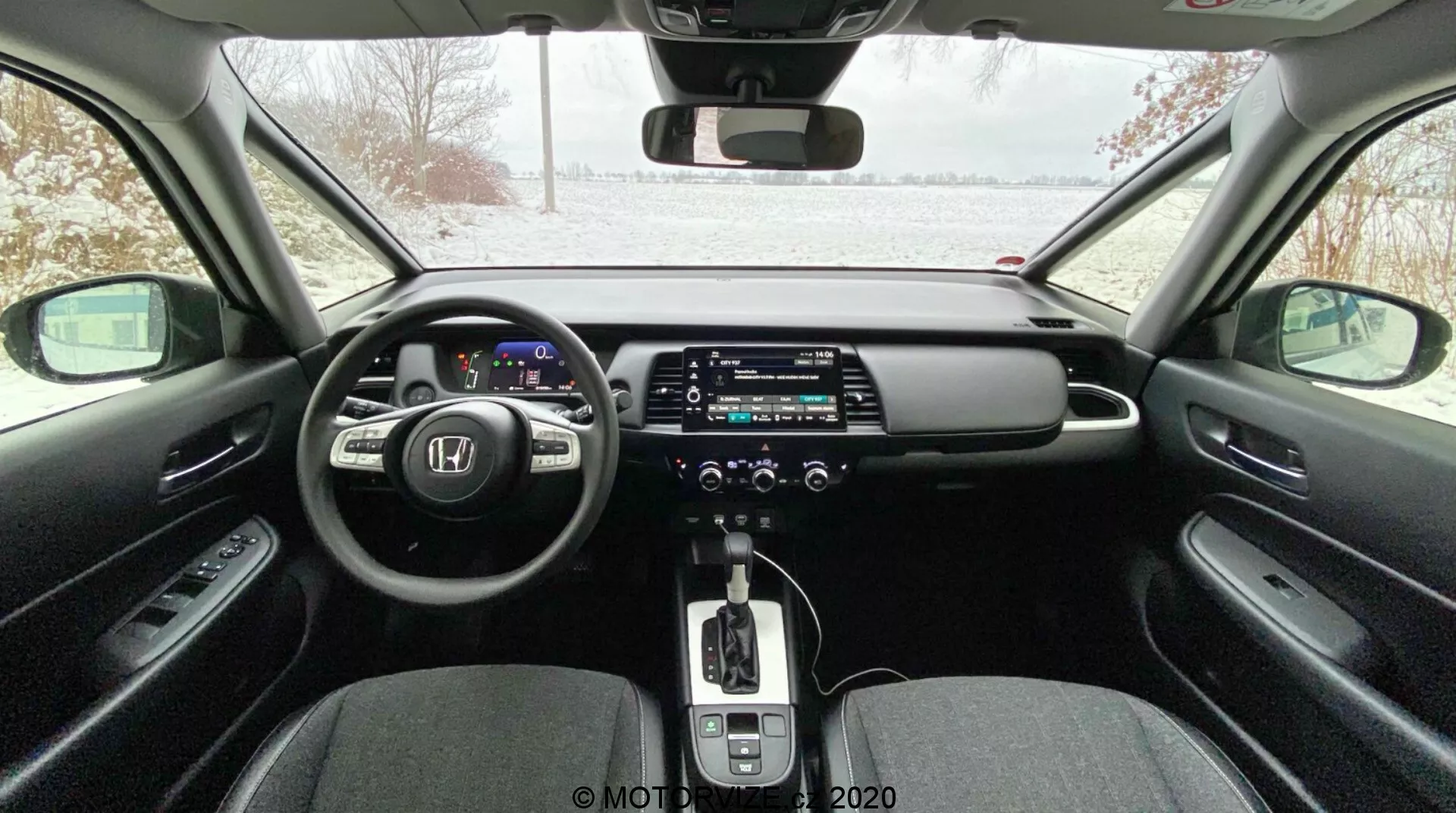 Moderný interiér auta vidieť zo zadného sedadla, obsahuje digitálny prístrojový panel, multifunkčný volant, veľký dotykový infotainment displej, automatickú prevodovku s koženou radiacou pákou, dvojzónovú klimatizáciu, elektronickú parkovaciu brzdu a elegantný dizajn s kontrastnými materiálmi; zasnežený exteriér viditeľný cez čelné sklo naznačuje možnú zimnú atmosféru.