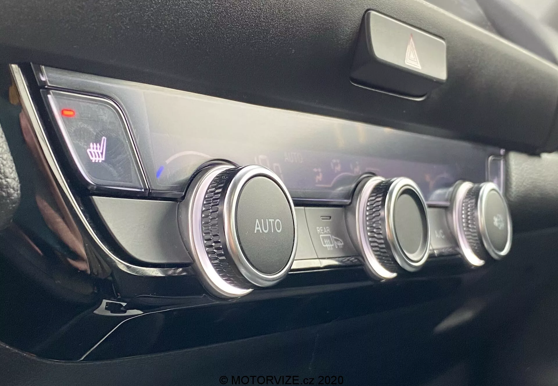 Detailný pohľad na moderný ovládací panel klimatizácie automobilu Honda Jazz 2020 s tromi kruhovými ovládačmi s kovovým povrchom, automatickým klimatizačným systémom, nastavením teploty v dvoch zónach indikovaným na digitálnom displeji a osvetlenými ikonami vyhrievania sedadiel, všetko zasadené v minimalistickom a kvalitnom dizajne prístrojovej dosky.