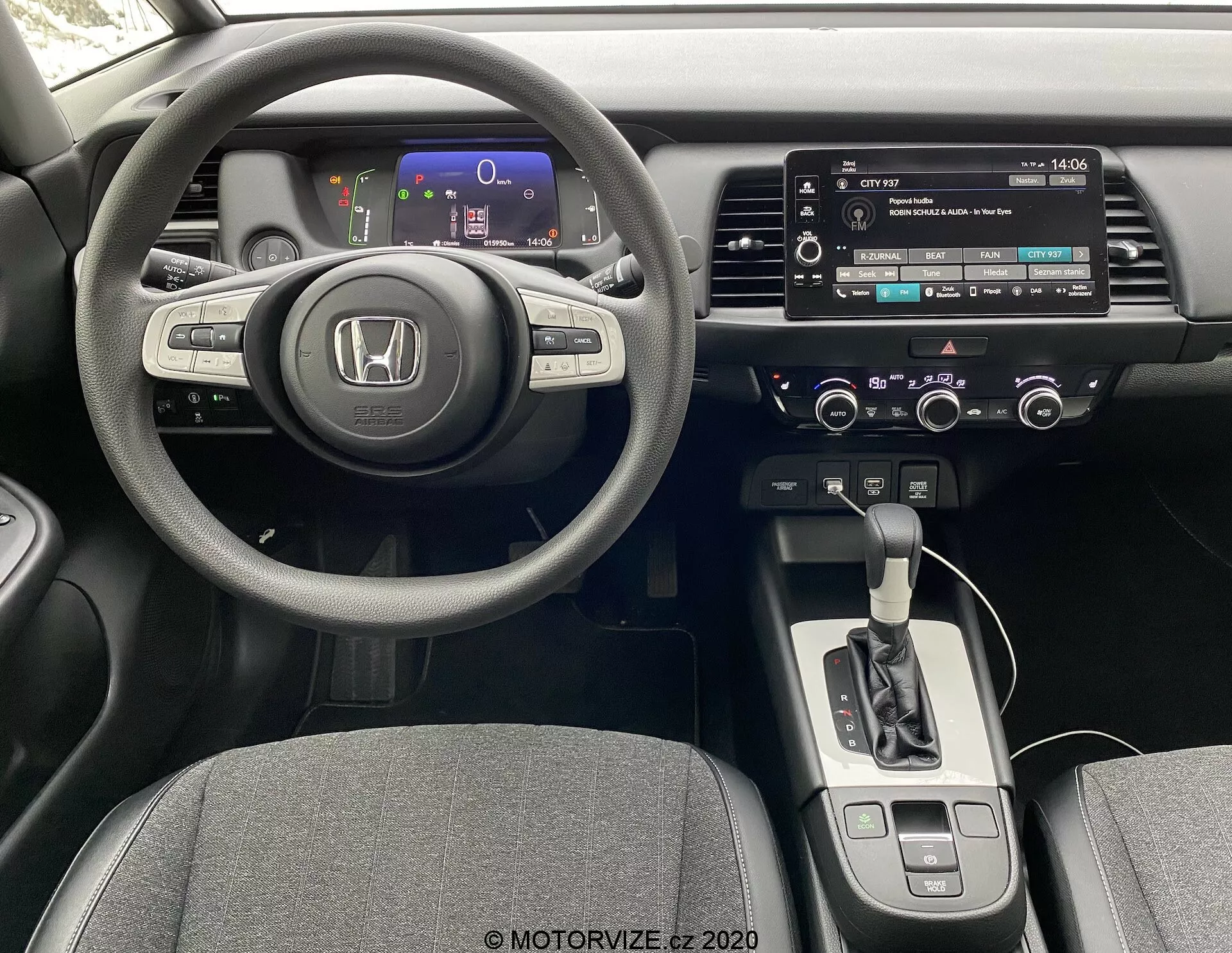 Interiér auta Honda Jazz 2020 z pohľadu vodiča, zobrazujúci sofistikovaný trojramenný volant s ovládacími prvkami, digitálny prístrojový panel s farebnými ukazovateľmi, centrálnu konzolu s dotykovým infotainment systémom, manuálne ovládanie audia a klimatizácie, automatický voliť prevodovky, a sedadlá čalúnené látkou.