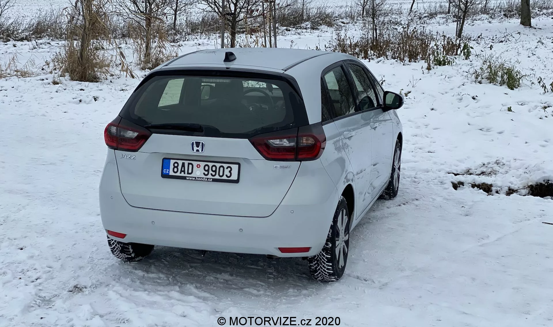 Obrázok zobrazuje biely hatchback Honda Jazz 2020 z pohľadu tri štvrtiny zozadu, stojaci na povrchu pokrytom snehom, s bezlistými stromami v zimnej scenérii a zatiahnutou oblohou v pozadí. Zadný dizajn zahŕňa zadný stierač čelného skla, emblém výrobcu, označenie modelu a zadné osvetlenie s červenými a priehľadnými šošovkami. Bočný profil ukazuje zadné dvere pre cestujúcich, predné dvere, bočné okná a viditeľný uzáver paliva. Auto je vybavené viac-lúčovými liatinovými kolesami, označujúcimi vzor pneumatík vhodný na sneh s miernym nánosom snehu. Zadná registračná značka naznačuje geografický pôvod. Svetlá výška je typická pre hatchbacky, bez viditeľnej koncovky výfuku či vonkajších doplnkov. Auto vyzerá čisto a v dobrom stave, s minimálnou špinou na zadnom nárazníku kvôli zasneženým cestám.