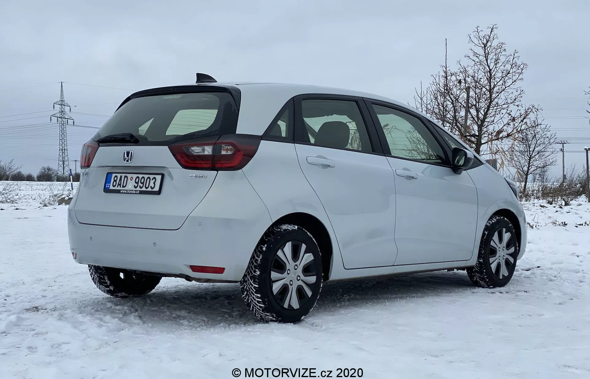Zadný trojštvrťový pohľad na hatchback Honda Jazz 2020 na snežnej ploche v predmestskom prostredí s holými stromami a elektrickými vežami v pozadí. Obrázok zachytáva pasažiersku stranu vozidla, moderný dizajn zadných svetiel pravdepodobne s LED technológiou, zadné dvere, zadný stierač, viditeľný zadný nárazník s integrovanými parkovacími senzormi a pravdepodobne zimné pneumatiky. Vozidlo je zaparkované v prirodzenom dennom svetle pod zamračenou oblohou, čo naznačuje zimné jazdné podmienky.