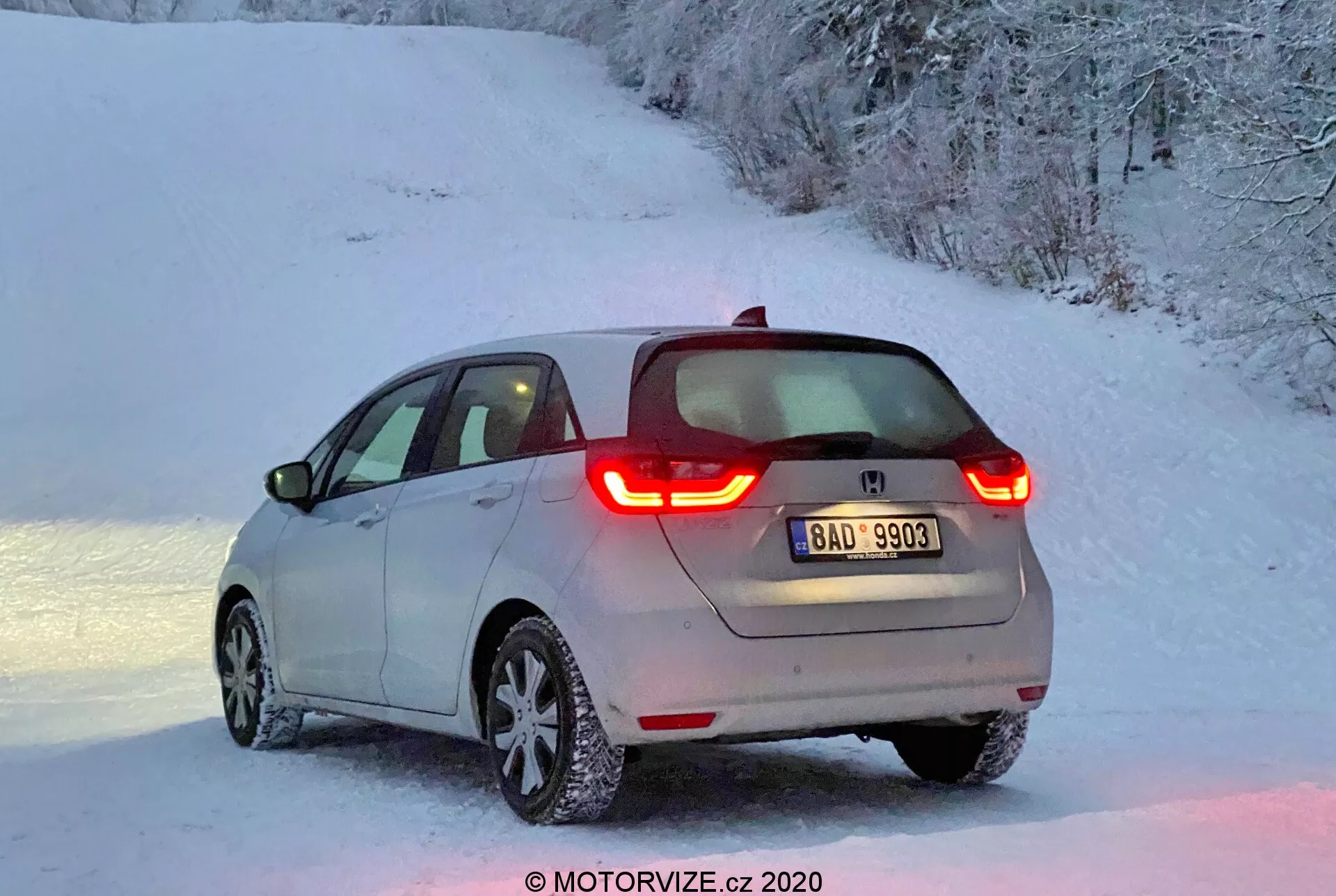 Moderný hatchback Honda Jazz 2020 v zadnom trojštvrťovom pohľade, osvetlený mäkkým svetlom úsvitu alebo súmraku, s výraznými osvetlenými C-tvarovanými LED zadnými svetlami, viditeľným logom výrobcu, ľahkým popraškom snehu na vozidle a zliatinovými kolesami so zasneženými pneumatikami v zasneženej vidieckej scenérii.