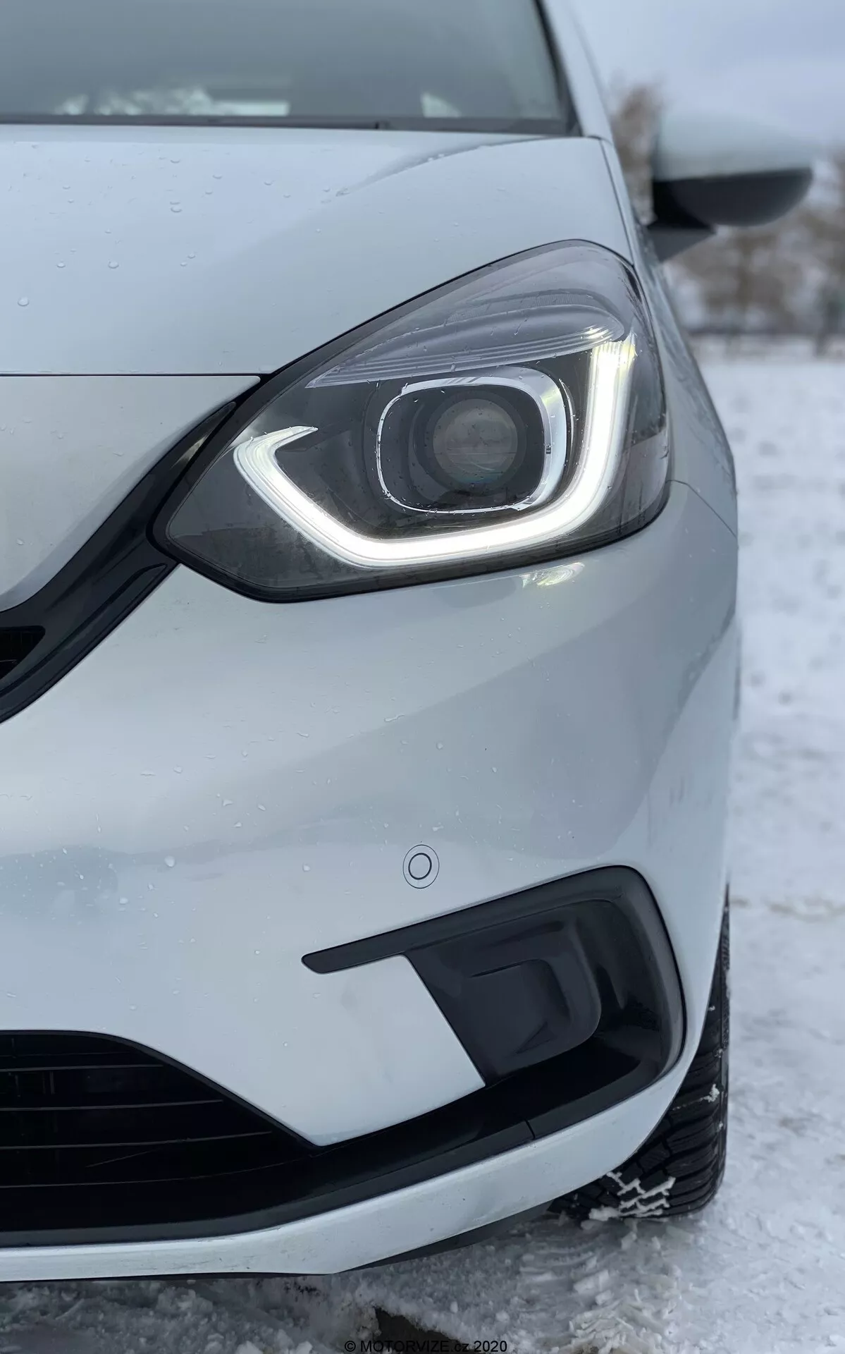 Detailný záber ľavej prednej strany vozidla Honda Jazz 2020, snímaný z nízkeho uhla. Zameriava sa na svetlomet s moderným LED dizajnom a dennými svetlami, ktoré sa ohýbajú okolo hornej a bočnej časti. Súčasťou svetlometu je viditeľná projekčná šošovka, čo naznačuje pokročilé HID alebo LED svetlomety pre hlavný lúč. Predný nárazník, zobrazený pod svetlometom, obsahuje mriežku prívodu vzduchu a kryt pre hmlové svetlo. Kovová strieborná alebo svetlo šedá farba automobilu a čiastočne viditeľné koleso a bočné zrkadlo sú podrobne popísané. Sneh a ľad na zemi naznačujú chladné podnebie, čo odráža výkon auta v zimných podmienkach. Obrázok tiež zvýrazňuje kvalitu stavby automobilu, medzery medzi panelmi a senzor blízkosti na nárazníku, ktorý je súčasťou parkovacej asistenčnej technológie.