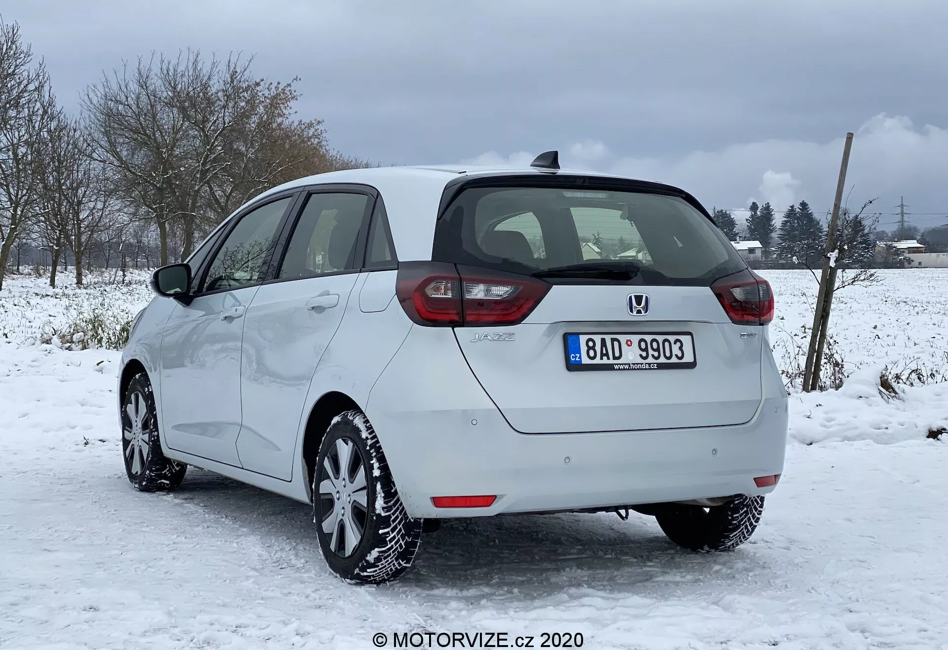 Obrázok strieborného hatchbacku Honda Jazz 2020 zaparkovaného v zasnežených podmienkach s vidieckym alebo polovidieckym pozadím. Zaznamenaný zozadu a z čiastočného bočného profilu z pravej strany, zvýrazňujúci vertikálne usporiadané zadné svetlá, centrálny znak a nárazník s parkovacími senzormi. Jasne viditeľné sú hladké bočné kontúry, hliníkové kolesá a zimné pneumatiky, spolu s funkciou odmrazovania okien a zadného skla a nezasúvajúce bočné zrkadlá vo farbe karosérie. Viditeľná je aj poznávacia značka, strešná anténa a celkový dobre udržiavaný stav vozidla.