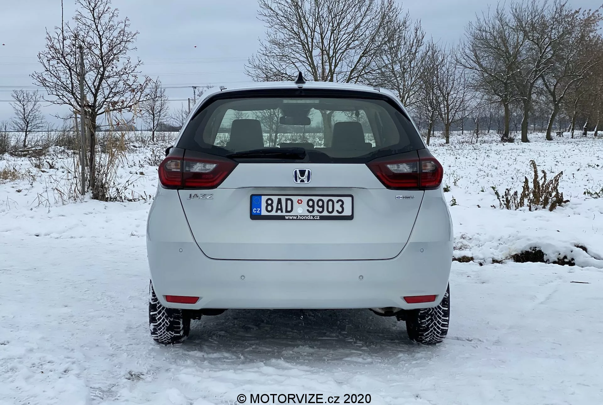 Zadný pohľad na strieborný hatchback Honda Jazz 2020 zaparkovaný na ľadovom povrchu. Obrázok jasne zachytáva zadný nárazník, zadné dvere, koncové svetlá a zadné okno vozidla. Na zadných dverách je viditeľný emblém výrobcu a názov modelu spolu so štandardnou evidenčnou značkou umiestnenou v strede. Auto má osvetlené osvetlenie evidenčnej značky, odrazky na oboch koncoch zadného nárazníka a koncové svetlá so štandardnou žiarovkou. Zadné okno má funkciu odmrazovania označenú vodorovnými čiarami a zadný stierač je zaparkovaný navrchu. Výfukový systém je mierne viditeľný pod zadným nárazníkom na pravej strane. Auto má oceľové kolesá s dekoratívnymi krytmi a nachádza sa v zimnom krajinnom prostredí so snehom a holými stromami v pozadí.