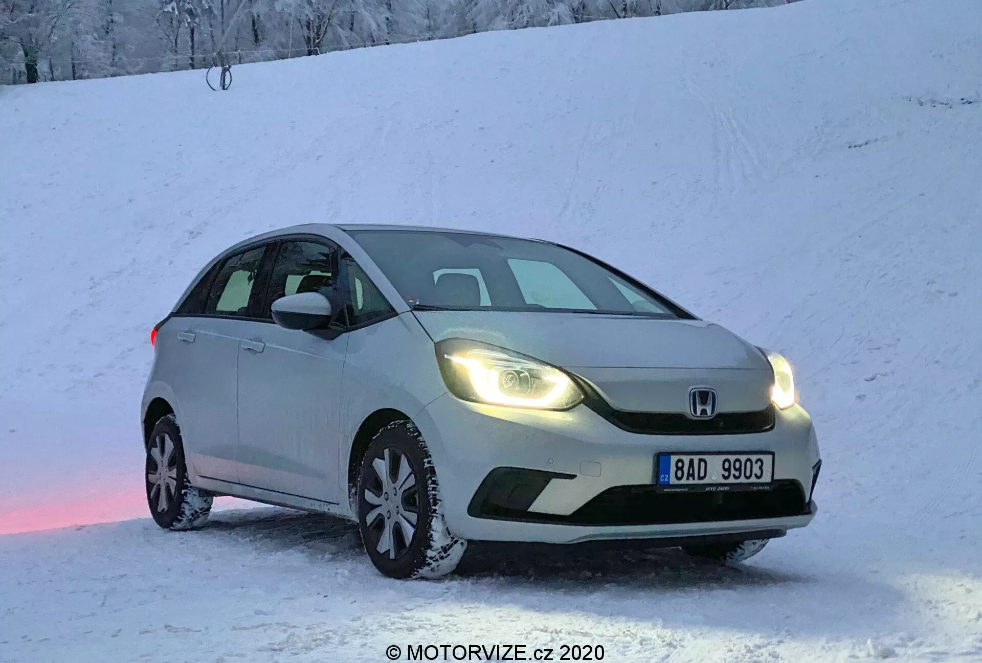 Strieborný hatchback Honda Jazz 2020 zaparkovaný na zasneženej ploche počas svitania alebo súmraku s teplými zapnutými svetlami, zachytený z predného trojštvrtinového pohľadu. Obrázok zvýrazňuje prednú masku, svetlomety, kapotu, čelné sklo a časť vodičovej strany. Viditeľná európska poznávacia značka, sneh na zemi a aute, vytvára chladnú, no útulnú atmosféru. Mierne ružové svetelné odrazy na snehu pod autom. Fotografia z nízkeho uhla zdôrazňuje vybavenie zimnými pneumatikami a dynamický vzhľad pre schopnosti riadenia v zime.