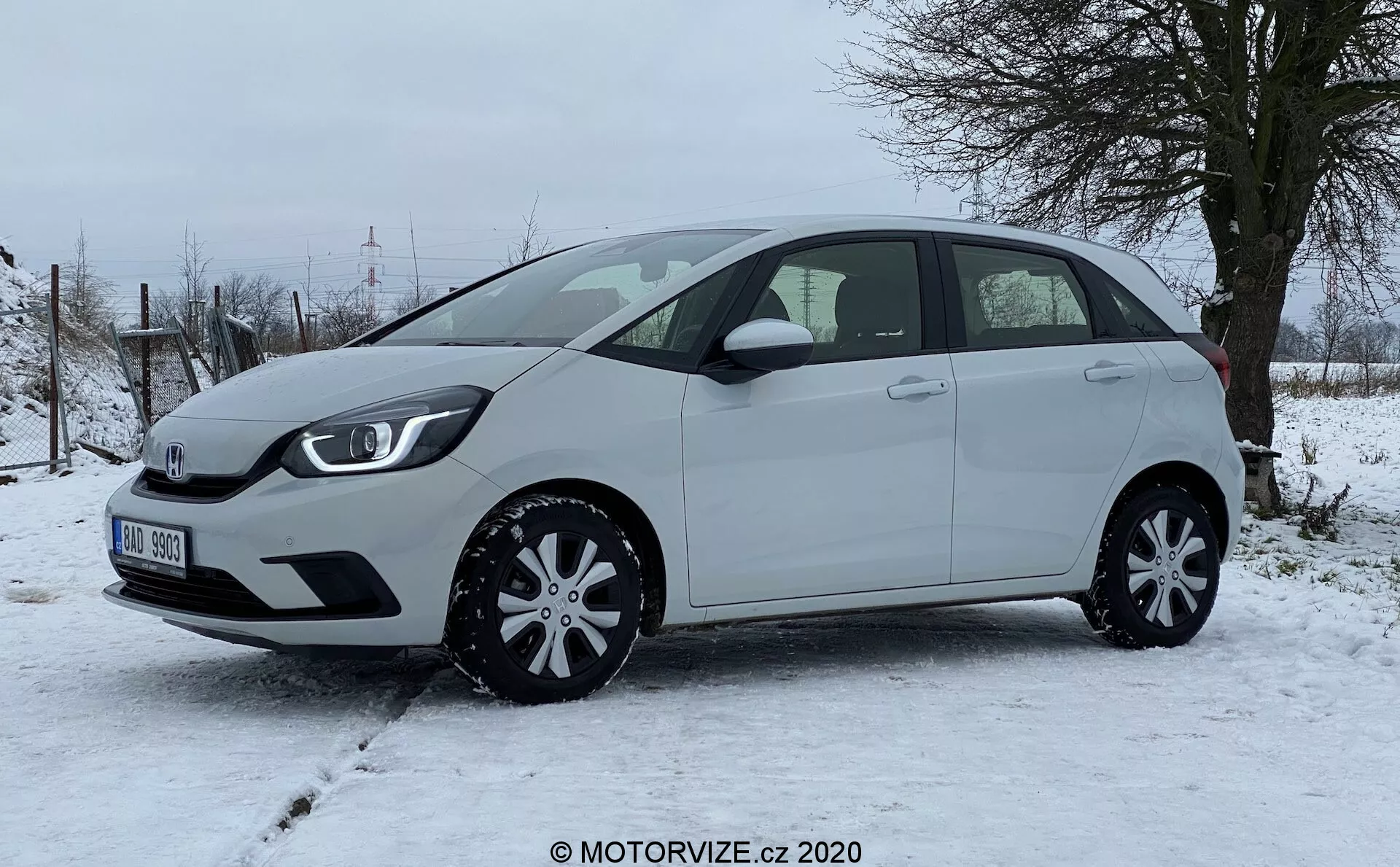 Moderný hatchback Honda Jazz 2020, fotografovaný z boku, zvýrazňujúci ľavý profil vozidla na zasneženej ploche s vidieckou vegetáciou v pozadí. Kompaktné vozidlo má mierne klesajúcu líniu strechy, päť dverí vrátane zadných výklopných, bočné zrkadlá v farbe karosérie a štýlové kryty kolies, vyžarujúce praktickosť a efektívnosť dizajnu. Z tohto uhla nie sú viditeľné žiadne pokročilé vonkajšie prvky, ako sú kamery alebo senzory, čo zdôrazňuje jasný pohľad na proporcie auta, kontúry dverí, líniu okien a oblúky kolies.