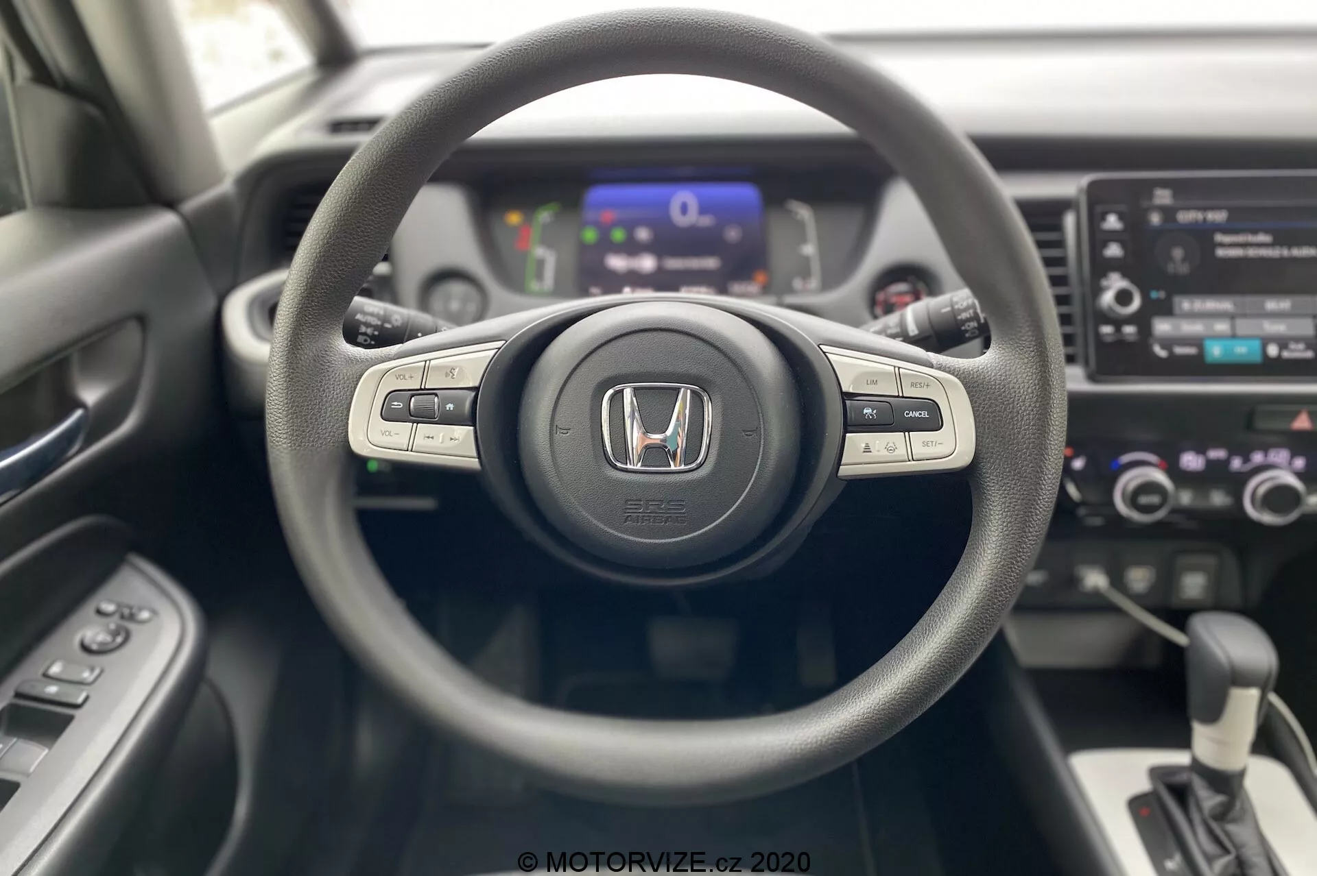 Interiér Honda Jazz 2020 z pohľadu vodiča, zvýrazňujúci stredovo umiestnený trojramenný volant s emblémom Honda, ovládacími tlačidlami pre médiá a tempomat na ramenách, digitálny prístrojový panel v mierne rozmazanom pozadí a stredovú konzolu s dotykovým infotainment systémom, dvojzónovú klimatizáciu a automatickú radiacu páku. Moderný a súčasný dizajn s rôznymi textúrami materiálov.