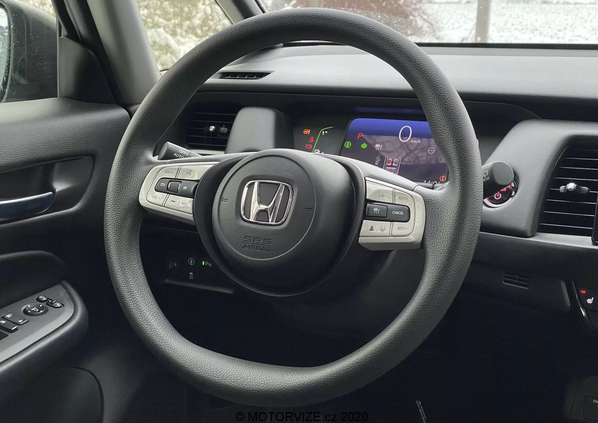 Pohľad na interiér Honda Jazz 2020 z perspektívy sedadla vodiča, zameraný na trojramenný volant s odznakom Honda a integrovanými ovládacími prvkami pre audio, tempomat a pravdepodobne Bluetooth alebo hlasové ovládanie. Za volantom je digitálny prístrojový panel s farebnými displejmi zahŕňajúcimi rýchlomer a otáčkomer. Čiastočne viditeľná je stredová konzola s infotainment alebo navigačnou obrazovkou a ventilačné otvory. Panel dverí vodiča s tlačidlami na ovládanie okien a bočných zrkadiel je tiež čiastočne viditeľný.