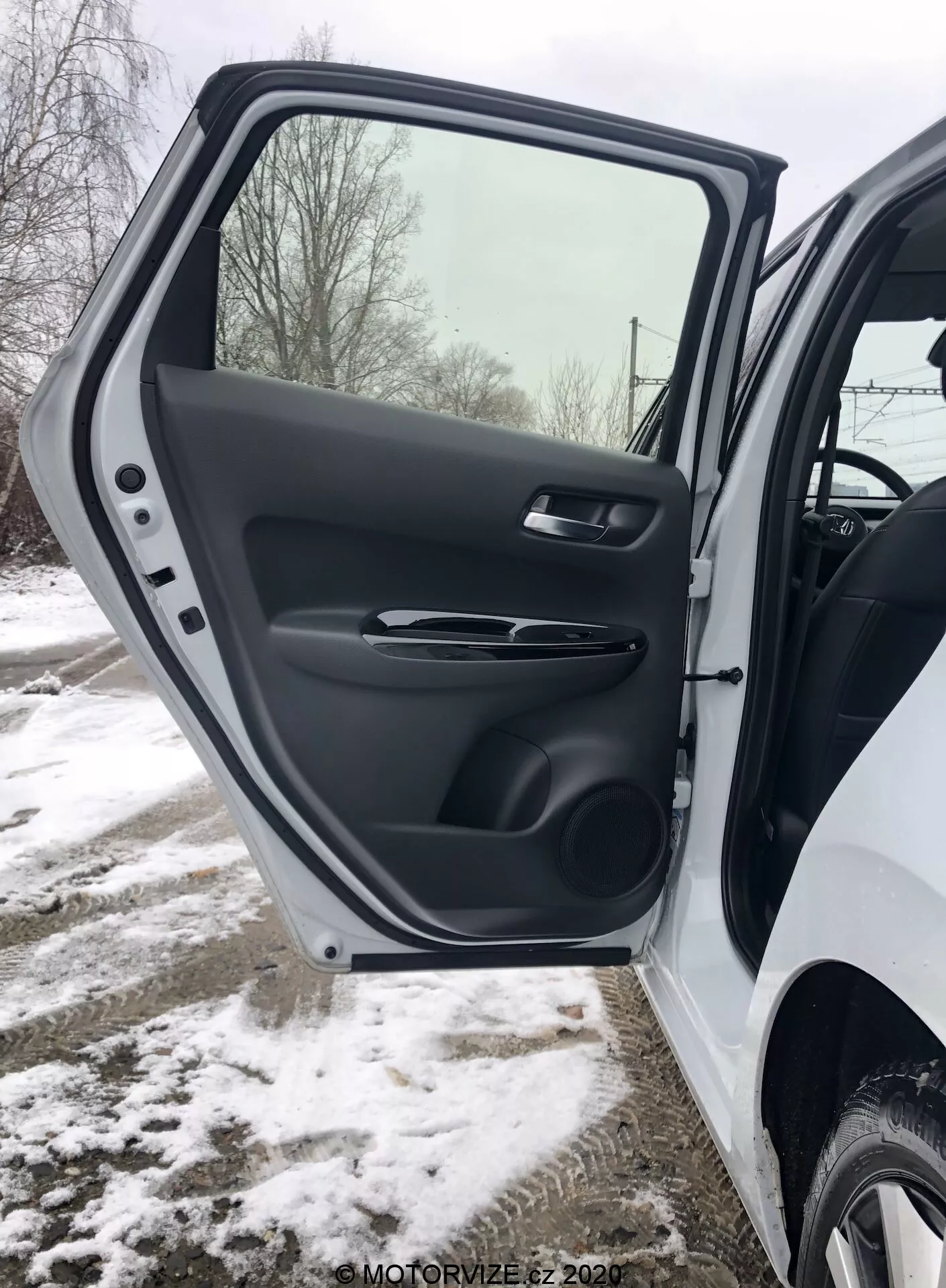 Detailný pohľad na interiér predných dverí vodiča Honda Jazz 2020, zobrazujúci textúrovaný plastový obklad, saténovo zakončenú ergonomickú kľučku, ovládanie elektrických okien a zrkadiel, tvarovanú úložnú kapsu a mriežku reproduktora, všetko v zasneženom zimnom prostredí.