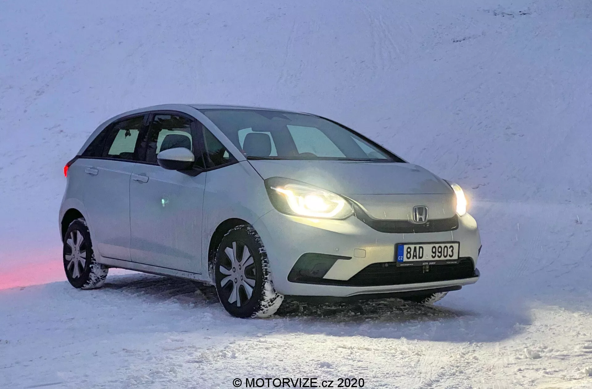 Predný trojštvrťový pohľad na svetlý hatchback Honda Jazz 2020, umiestnený na zasneženom povrchu. Obrázok zachytáva auto z prednej ľavej strany, pričom ukazuje stranu vodiča v uhle, ktorý zvýrazňuje bočný profil aj prednú časť. Honda Jazz je zaparkovaná s predkom mierne natočeným doprava, čo odhaľuje dizajn mriežky chladiča, svetlometov a časti kapoty. Predné svetlá sú zapnuté a odrážajú sa od snehu, auto pôsobí staticky. Viditeľné sú zimné pneumatiky s výraznou dezénovou vzorkou, ktoré zdôrazňujú vhodnosť pre zimnú jazdu.