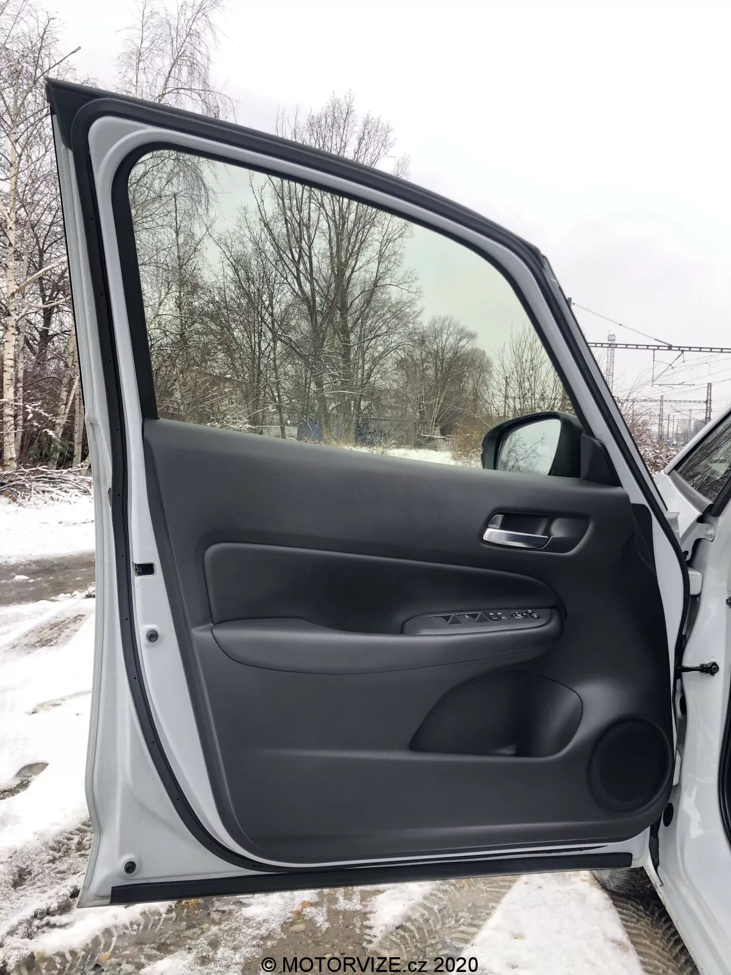 Interiérový pohľad na predné dvere spolujazdca Honda Jazz 2020, úplne otvorené, zobrazujúce textúrované a hladké čierne plastové materiály. Panel dverí zahŕňa ergonomickú opierku, mäkkú rukoväť a ovládací panel pre elektrické ovládanie okien a zamykacie mechanizmy. Viditeľná je aj strieborná lišta a spodné úložné vrecko. Cez okno je vidieť zasnežená krajina, čo naznačuje zimné prostredie.