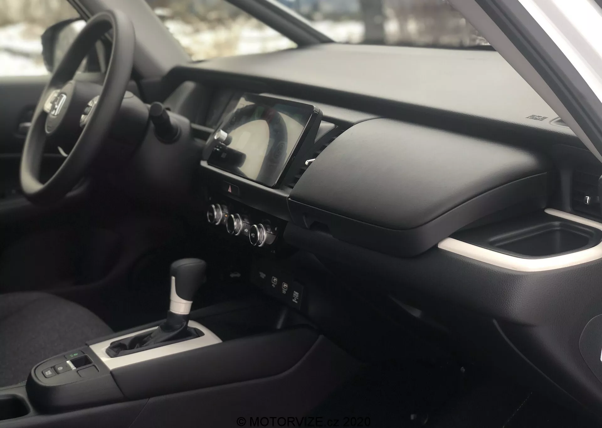 Interiérový pohľad na Honda Jazz 2020 z pohľadu spolujazdca, zameraný na prístrojovú dosku a kokpit vodiča. Fotografia, zhotovená pod mierne zvýšeným uhlom, zvýrazňuje centrálnu konzolu, multifunkčný volant s ovládacími prvkami, digitálno-analógový prístrojový panel, stredovú konzolu s radiacou pákou, ovládacie gombíky klimatizácie a infotainment/navigačnú obrazovku. Podrúčka naznačuje pohodlie vodiča a interiér obsahuje kovové a soft-touch materiály, ktoré odrážajú modernú a vysokokvalitnú koncepciu dizajnu.