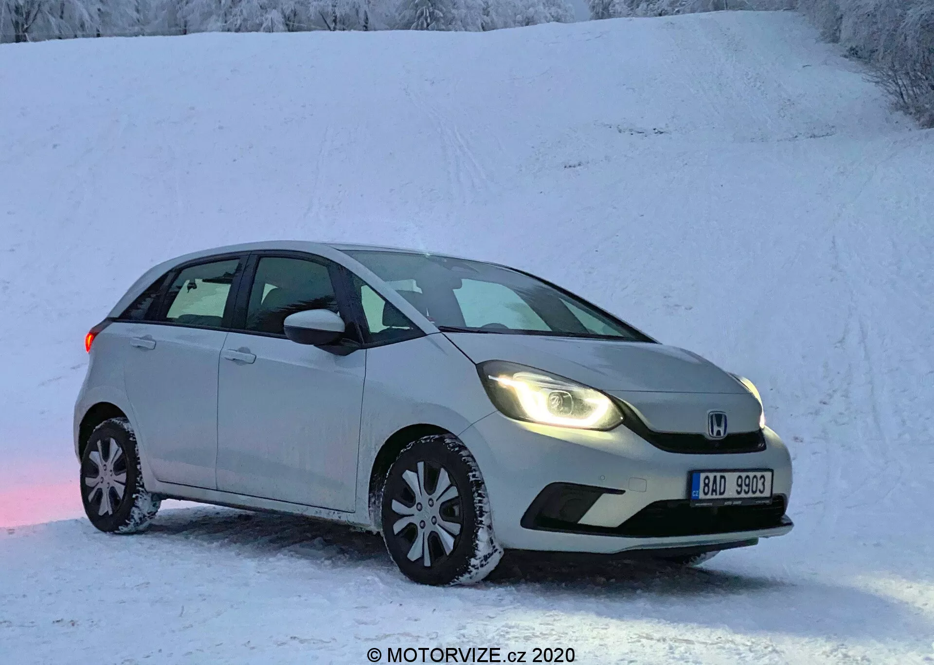 Obrázok predstavuje Honda Jazz 2020, strieborný alebo svetlošedý hatchback stojaci na zasneženej ploche, naznačujúci zimnú scénu so zasneženou krajinou. Zachytený z trojštvrťového predného uhla, výhľad zdôrazňuje vodičovu stranu, ukazujúc prednú mriežku, svetlomety, kapotu, čelné sklo, dvere vodiča a zadné dvere spolujazdca. Auto má moderné svetlomety, tvarovaný predný nárazník, prominentný znak na mriežke, bočné zrkadlá s integrovanými smerovými signálmi a viacramenné zliatinové kolesá s zimnými pneumatikami. Stacionárne vozidlo je zobrazené bez rozmazania pohybu za možných nízkych svetelných podmienok, buď skoro ráno alebo neskoro popoludní.