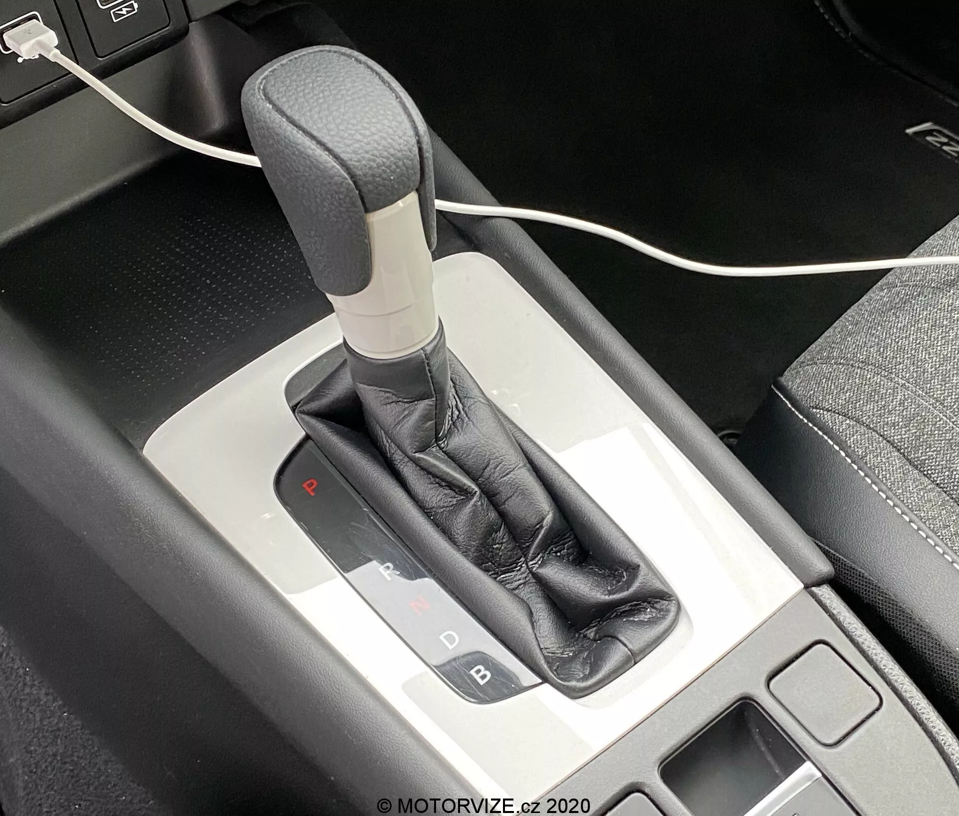 Detailný pohľad na automatickú radiacu páku Honda Jazz 2020 z pohľadu zhora, zobrazujúci mechanizmus voliča prevodových stupňov a okolitú stredovú konzolu. Radiaca páka má sivú alebo striebornú rukoväť, obalenú čiernym koženým obalom a doplnenou kovovým lemovaním. Osvetlený panel voliča prevodoviek ukazuje, že vozidlo je vo parkovacom režime s červeným svetlom pod 'P'. Na ľavej strane je vidieť malý úložný priestor s čiernou podložkou a v hornej časti obrázka čiastočne viditeľný USB kábel.