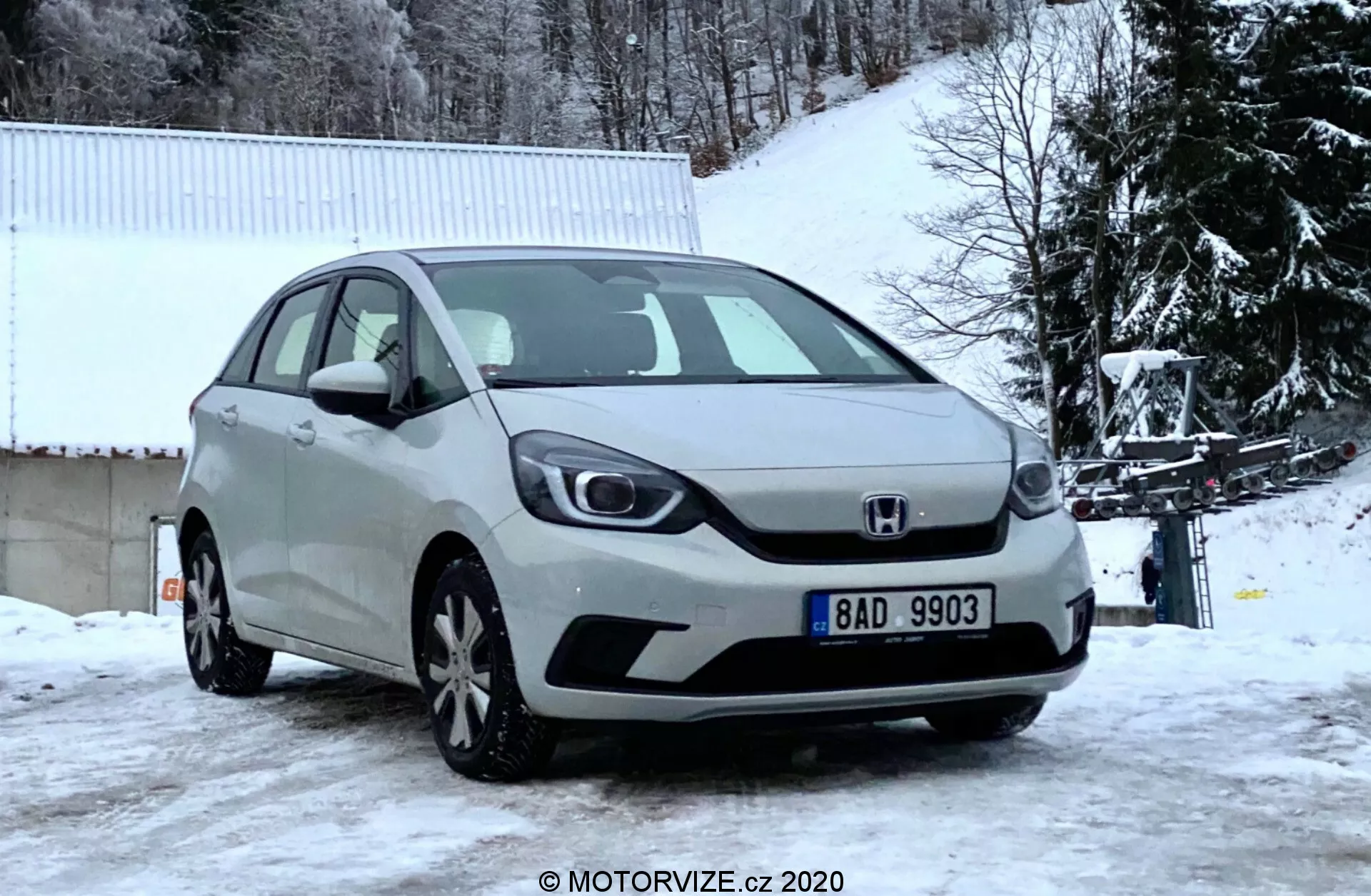 Predný uhol pohľadu na strieborný Honda Jazz hatchback z roku 2020 zachytený v zimnom dni, zobrazujúci prednú mriežku, svetlomety, kapotu a predný nárazník na pozadí zasneženej krajiny so stromami bez listov a bielou budovou.