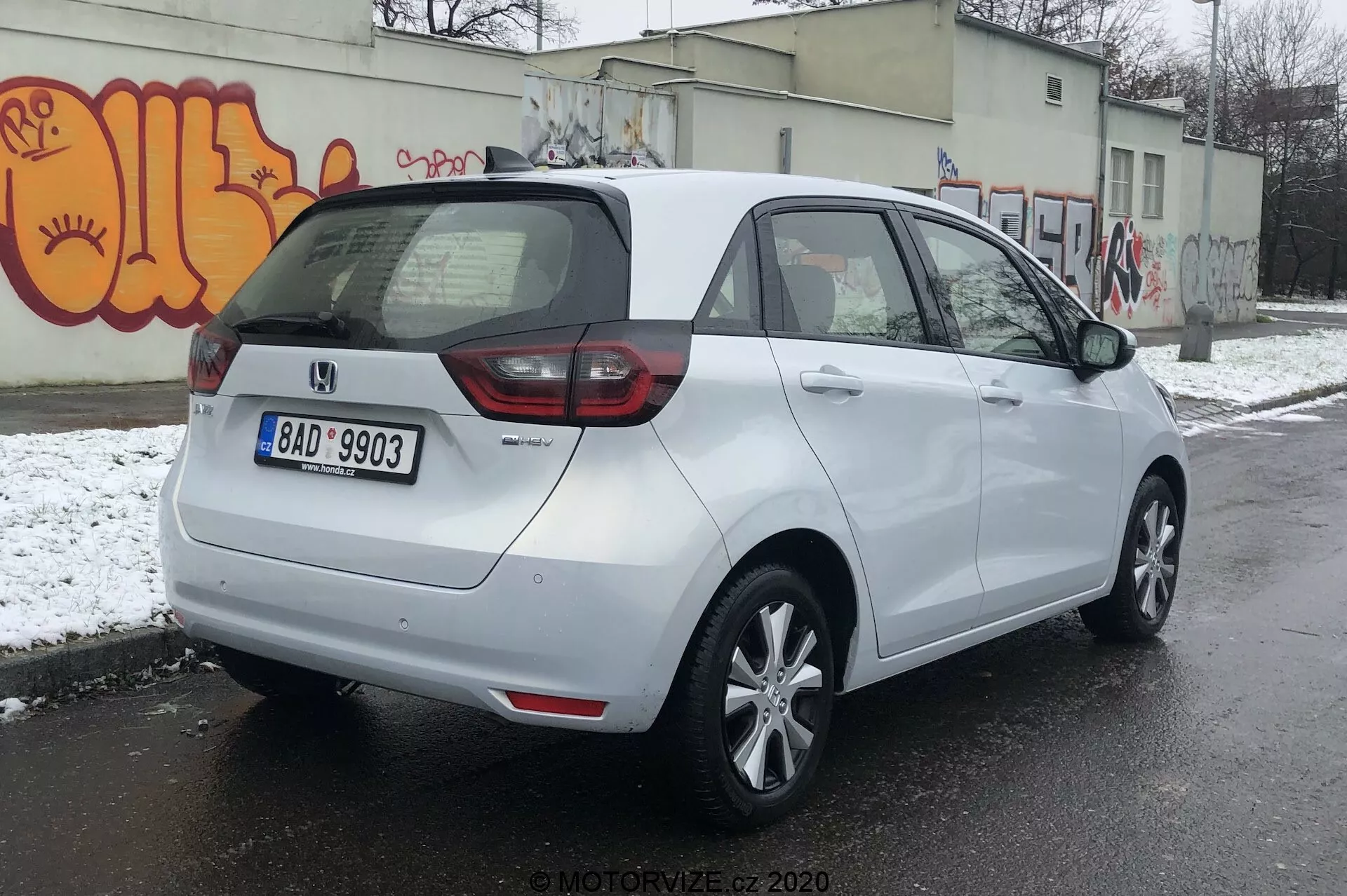 Trojštvrťový zadný pohľad na biely hatchback Honda Jazz 2020 zaparkovaný na asfaltovom povrchu v potenciálne chladnom mestskom prostredí snehom na zemi a graffiti na stene. Auto má mix zadných svetiel LED a tradičných žiaroviek, centrálnu zadnú ŠPZ a odznak naznačujúci úroveň výbavy alebo variant. Má aerodynamickú strechu, päťlúčové zliatinové kolesá, bokom lakované zrkadlá s integrovanými smerovkami a lakované kľučky dverí. Zadné okno je vybavené odmrazovačom a stieračom pre lepšiu viditeľnosť.