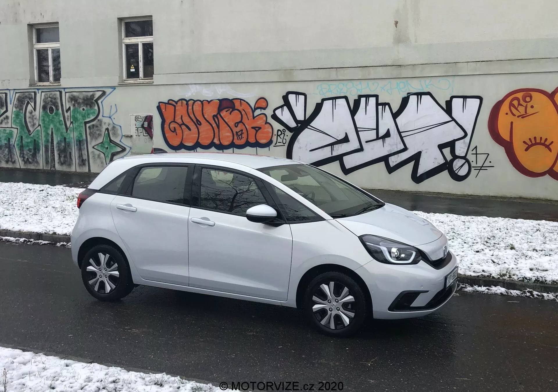 Obrázok predstavuje biely hatchback Honda Jazz 2020 zaparkovaný kolmo ku kamere na mestskej ulici, čo umožňuje celkový bočný pohľad. Čiastočne viditeľná predná časť vozidla s modernými svetlometmi prechádza do jemnej línie na boku. Kľučky dverí, bočné zrkadlá v rovnakej farbe ako karoséria a viacramenné kryty kolies zvyšujú jeho elegantný vzhľad. Priestranné presklenie ponúka dostatok miesta pre hlavu a zadné štvrťové okno prispieva k jeho dizajnu. Zadný spojler naznačuje aerodynamickú účinnosť. Zamračené osvetlenie s čerstvým snehom a mestská grafiti kulisa vytvárajú dynamické prostredie.