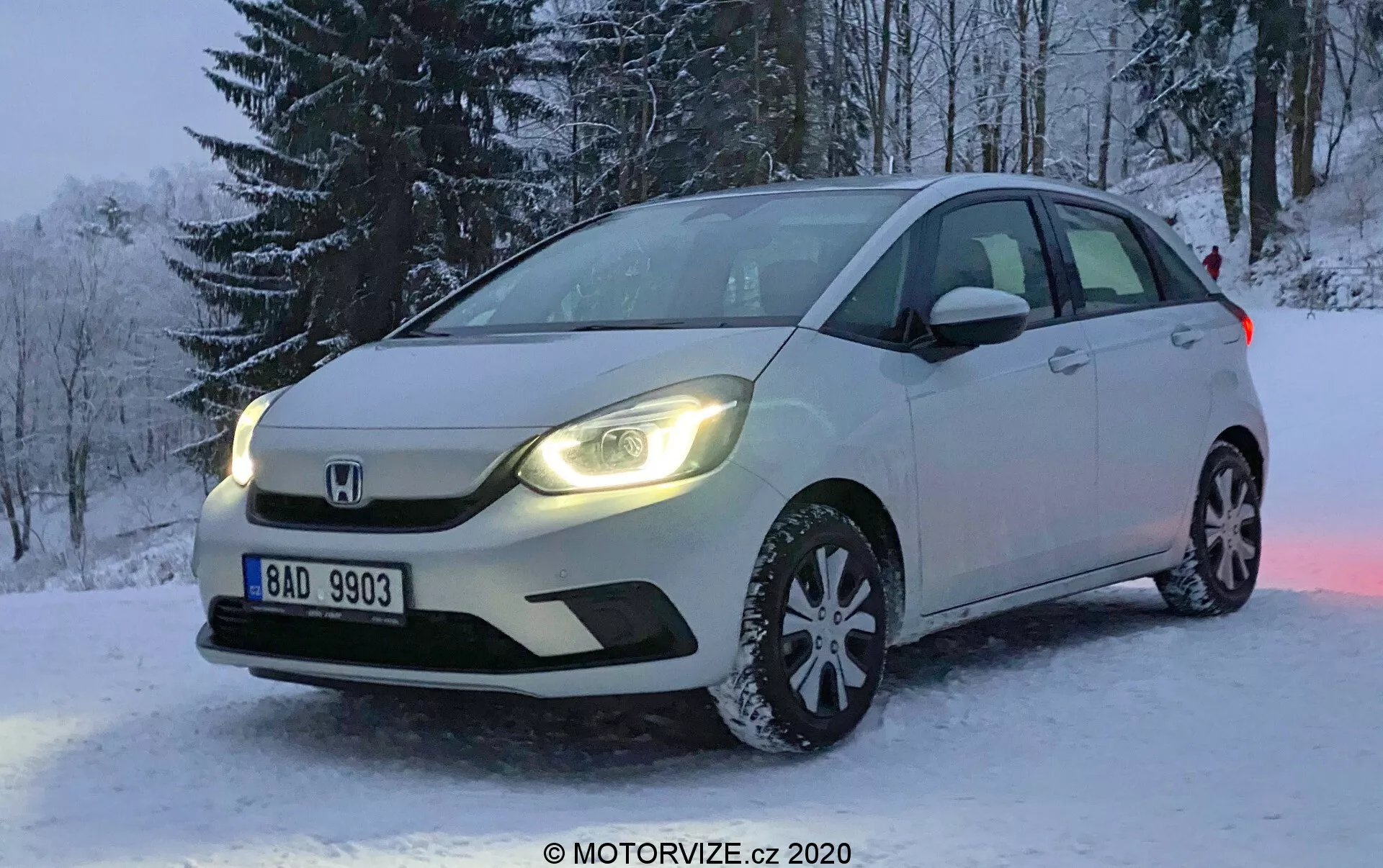 Profesionálna fotografia bieleho hatchbacku Honda Jazz 2020 zaparkovaného na zasneženom teréne počas zimy. Obrázok je urobený z uhla, ktorý sa zameriava na prednú ľavú stranu vozidla, ukazujúc prednú mriežku, LED svetlomety, kapotu, ľavé predné koleso vybavené snehovými pneumatikami, čelné sklo a dvere vodiča. V pozadí sú viditeľné stromy pokryté snehom a evidenčné číslo je čitateľné. Slabé svetlo naznačuje skoré ráno alebo neskoré popoludnie, s dennými svetlami vozidla odrážajúcimi sa od snehu a zdôrazňujúcimi scénu. Vozidlo má pokročilé osvetlenie a integrované smerovky v bočných zrkadlách, s výrazným znakom Honda na mriežke.