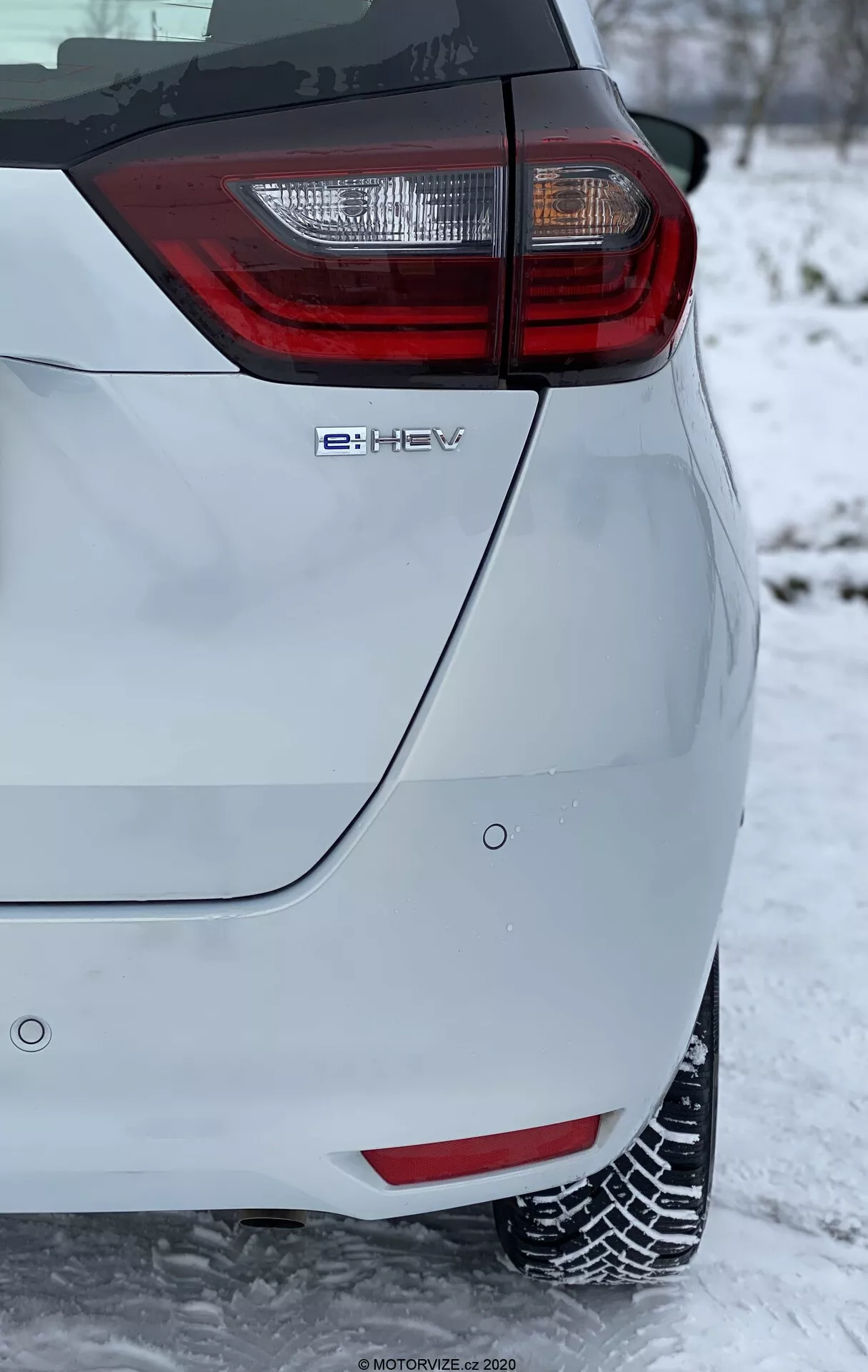 Detailný záber na zadný roh moderného hatchbacku, ktorý má výrazný zadný svetelný klaster s červenými a priehľadnými prvkami. Obrázok, zhotovený vo vertikálnej orientácii, ukazuje emblém hybridnej technológie 'e-HEV' a je zasadený do zimného prostredia so snehom na zemi a pokrývajúcim pneumatiky, čo naznačuje zimné jazdné podmienky. Svetlý lak vozidla kontrastuje so zasneženým pozadím, čím zvýrazňuje jeho moderné a ekologické dizajnové prvky.