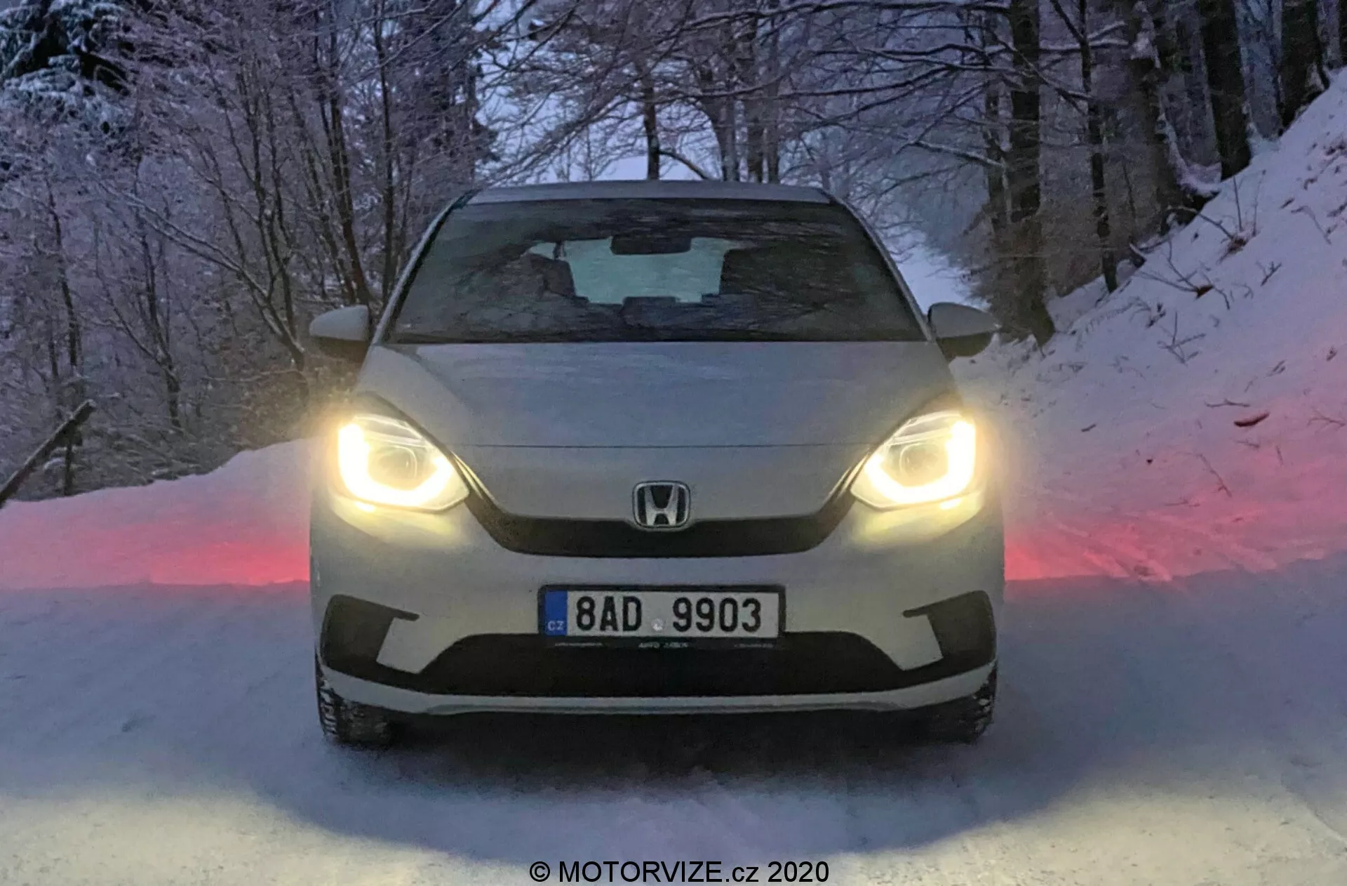 Čelný záber na Honda Jazz 2020 s LED svetlami, ukazujúc znak značky a EČ na mriežke, so sošným tvarom kapoty a tónovaným čelným sklom. Sneh na zemi a súmraková atmosféra zvýrazňujú pokročilý dizajn aj schopnosti vozidla v zimných podmienkach.