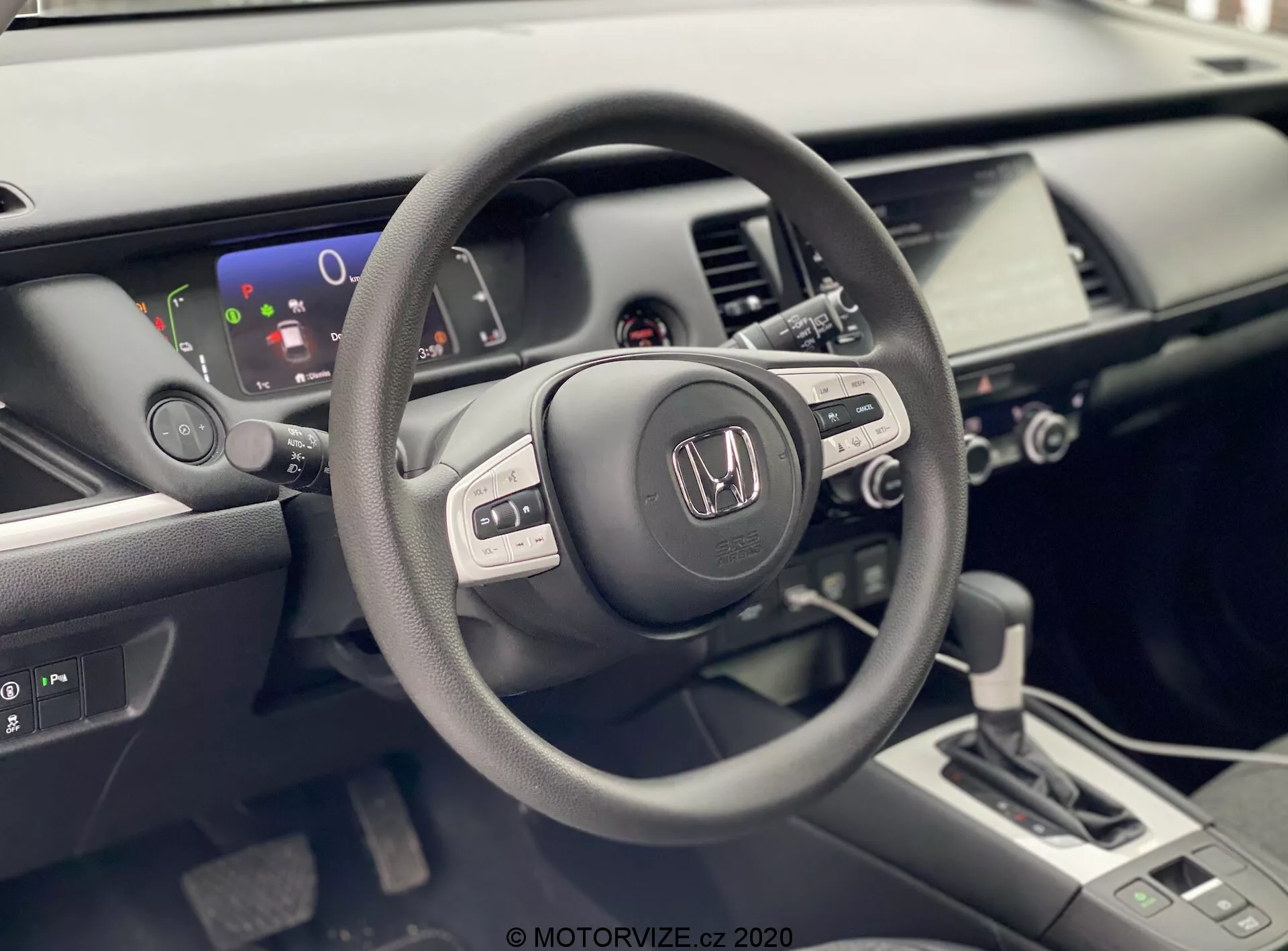 Fotografia zobrazuje interiér Hondy Jazz 2020 z pohľadu vodiča. Zameriava sa na volant s logom Honda, ktorý obsahuje viacero ovládacích tlačidiel naznačujúcich integrované rádio, informačný displej a možno aj tempomat. Za volantom je viditeľný čiastočný digitálny displej s ukazovateľom úrovne batérie, čo naznačuje, že vozidlo môže byť hybridné alebo plne elektrické. Dobre osvetlený, moderný interiér obsahuje aj stredový panel s dotykovým infotainment systémom, ovládaním klimatizácie a modernou pákou automatickej prevodovky. Obrázok zdôrazňuje matné, saténové a lesklé povrchy použitých materiálov interiéru, čo zvýrazňuje technologicky bohatý a ergonomický dizajn vozidla.