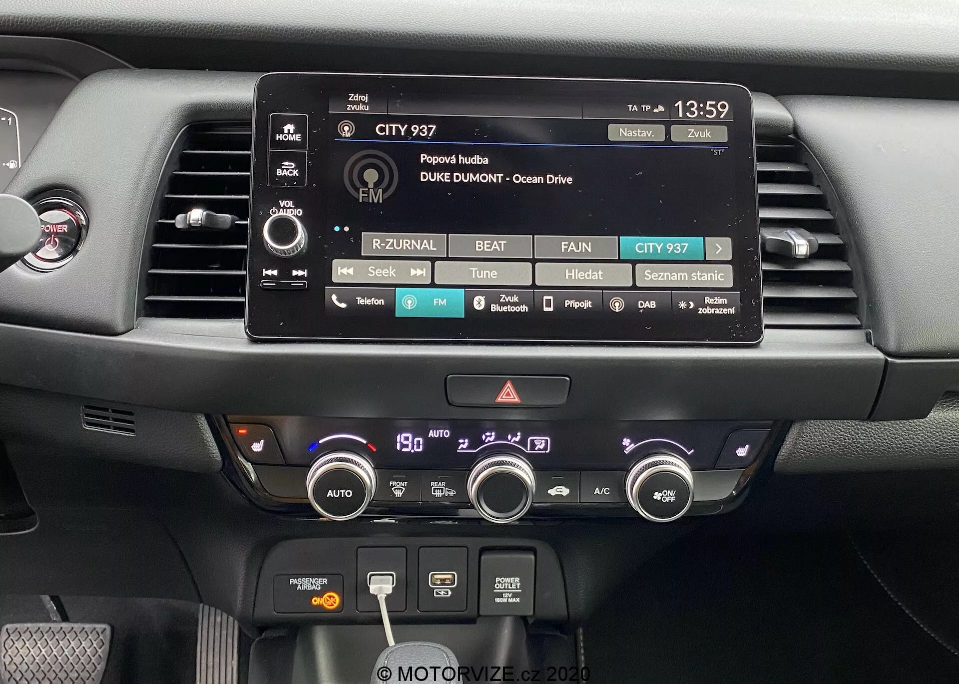 Detailný pohľad na centrálnu palubnú dosku modelu Honda Jazz 2020. Obrázok výrazne zobrazuje dotykový displej infotainmentu, obklopený fyzickými tlačidlami a otočnými gombíkmi pre nastavenie zvuku, médií a vozidla. Pod ním sú viditeľné dva vzduchové výduchy a komplexný klimatizačný systém s otočnými gombíkmi a tlačidlami na nastavenie teploty, rýchlosti ventilátora, odmrazovacie funkcie a recirkuláciu. Palubná doska je vyrobená z moderných materiálov vrátane matných a lesklých plastov, navrhnutá s ohľadom na funkčnosť a jednoduché používanie.