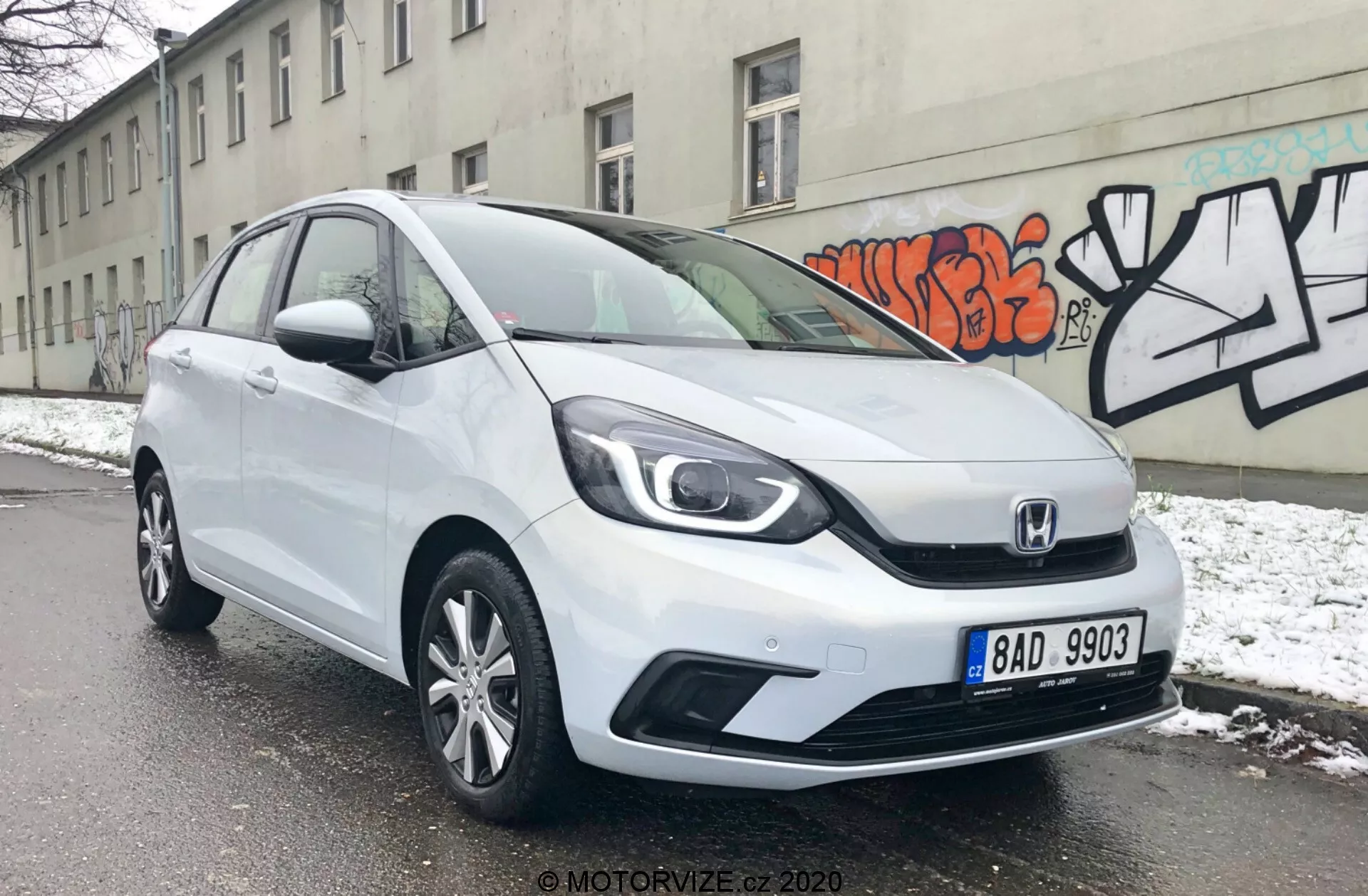 Obrázok ukazuje biele hatchback vozidlo Honda Jazz 2020 z čelného trojštvrťového uhla, odhaľujúc jeho elegantné svetlomety, charakteristickú dvojprúdovú mriežku Honda a aerodynamické kontúrové línie na kapote. Predný nárazník má viditeľné prívody vzduchu a pravdepodobne hmlové svetlá, zatiaľ čo bočný profil ukazuje kolesá z ľahkej zliatiny s viacerými lúčmi a mierne tvarované dverové panely. Auto je zaparkované na zasneženej ulici, čo naznačuje zimné prostredie, s pozadím šedej steny s graffiti, čo zvýrazňuje mestské prostredie.