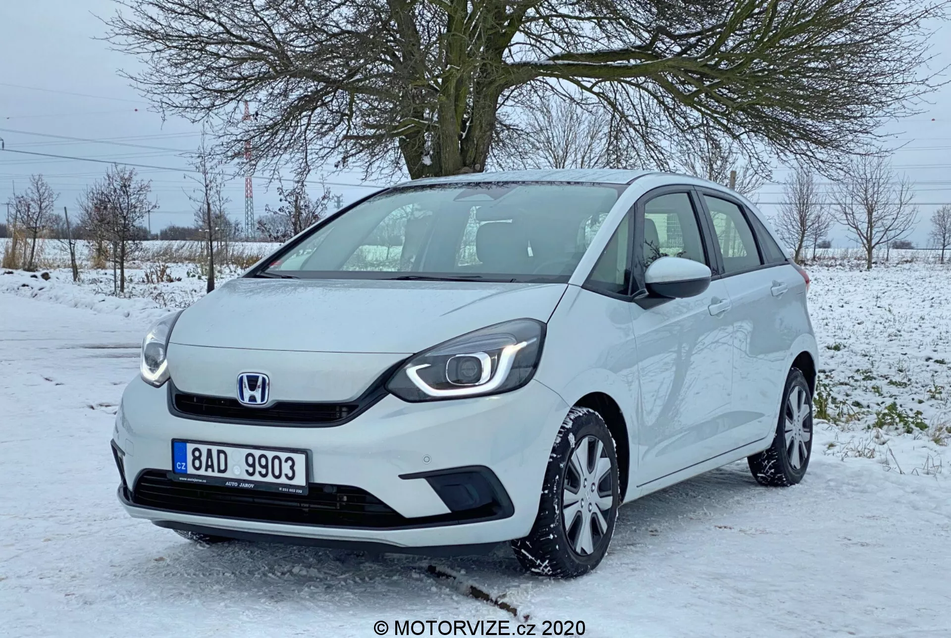 Pohľad na Hondu Jazz 2020 z predného trojštvrtinového uhla, ktorý ukazuje prednú časť a časť strany spolujazdca tohto moderného kompaktného hatchbacku. Obrázok, zrejme urobený z úrovne očí, zdôrazňuje minimalistickú mriežku s centrálnym emblémom Honda, elegantné svetlomety s LED dennými svetlami, viacramenné zliatinové kolesá a bočné zrkadlá s integrovanými smerovkami. Auto je zaparkované na čiastočne zasneženom spevnenom mieste, čo naznačuje zimné prostredie s lístia zbavenými stromami a šedou oblohou v pozadí. Vozidlo vyzerá čisto a dobre udržiavané.